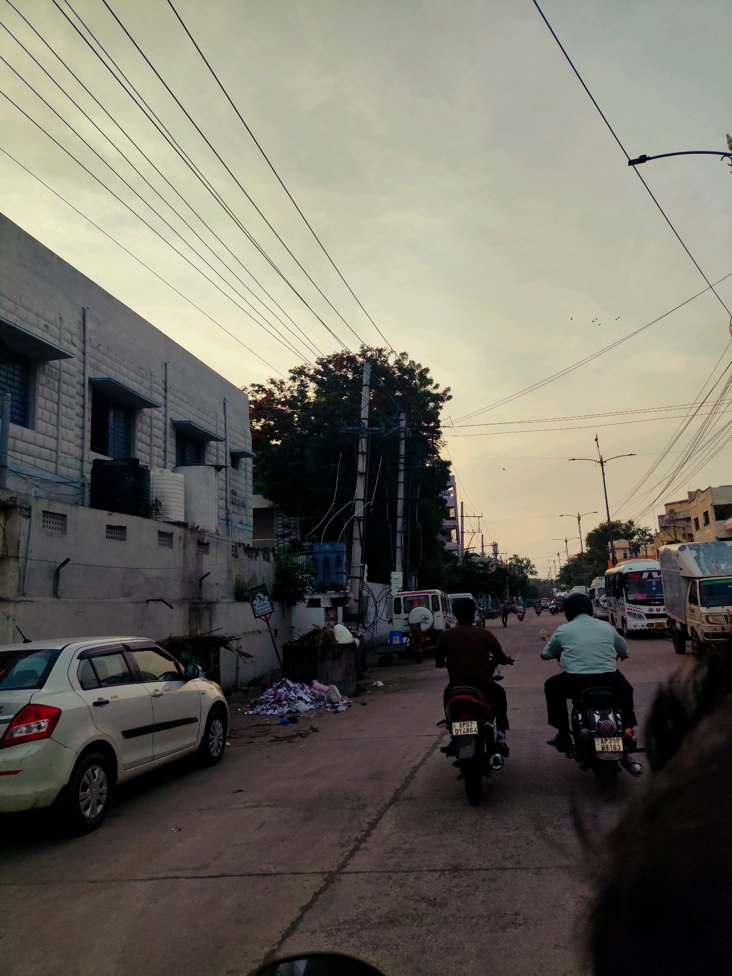 The image depicts a street scene in Guntur, India, under a hazy sky. In the foreground, a white sedan is parked on the left side of the road, facing away from the viewer. A pile of garbage is visible on the right side of the road, next to a dumpster. Further down the street, two individuals on motorcycles are riding away from the camera. The motorcycle on the left has a license plate that reads "AP07 DT4864." The motorcycle on the right has a license plate that reads "AP39R W8163." Several other vehicles, including buses and trucks, are present in the background, suggesting a commercial or mixed-use area. Buildings line both sides of the street, with some featuring balconies and windows. Overhead, a complex network of power lines crisscrosses the sky. The overall impression is one of a typical urban street during daylight hours, with the presence of traffic, buildings, and everyday urban elements like parked cars and garbage.
