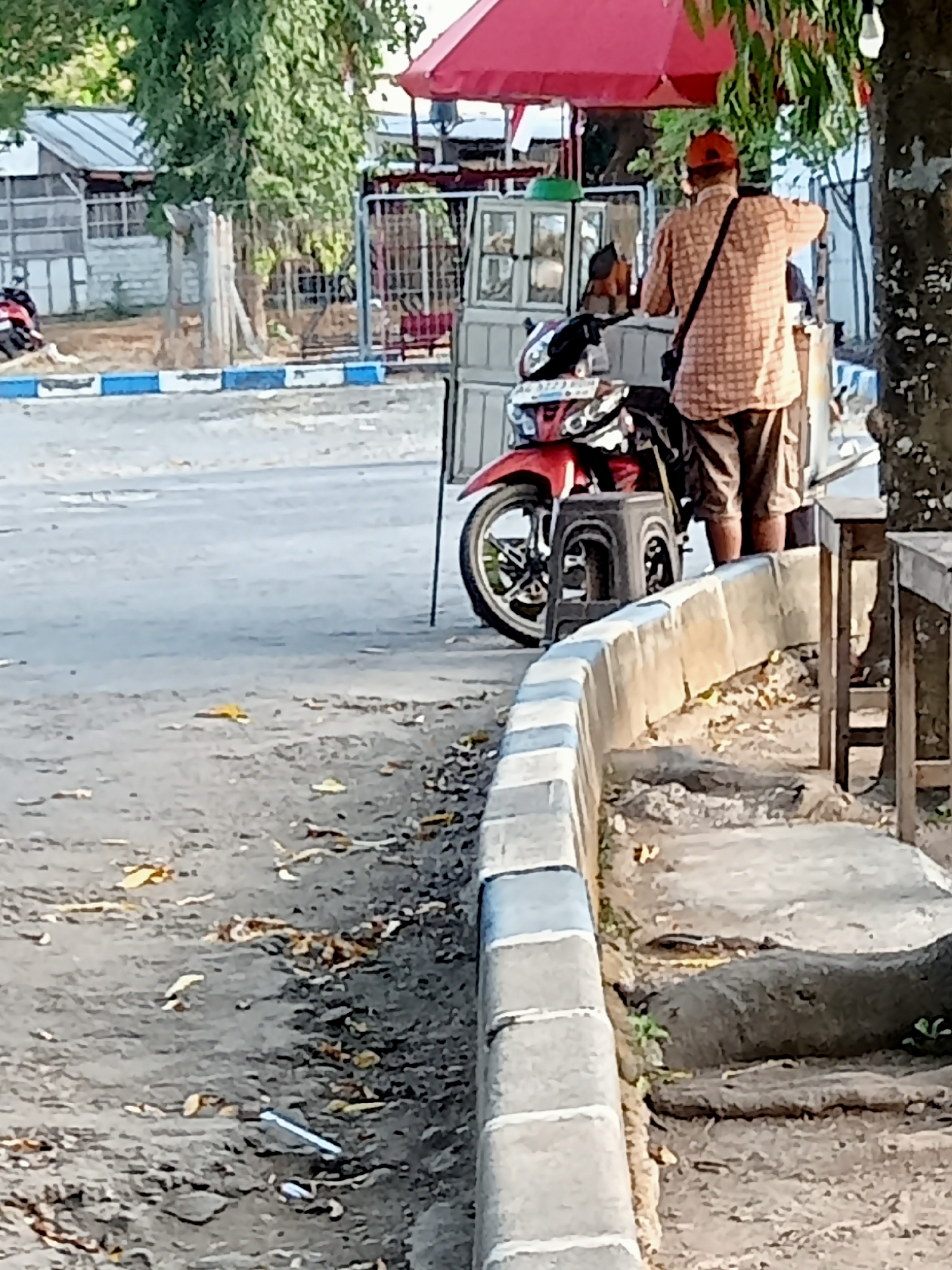 The image depicts a street scene in Majan, Indonesia, with a focus on a vendor's stall and a red motorcycle. A man wearing a checkered shirt and shorts stands behind the stall, his back to the camera. The stall appears to be mobile, equipped with a red umbrella and what looks like a dispenser or cooler. The red motorcycle is parked next to the stall, with its front wheel visible and resting on a black stool. The ground is paved with asphalt and a raised concrete curb lines the edge of the road. Fallen leaves are scattered on the ground, indicating an outdoor environment. In the background, a building with a tiled roof and a fence can be observed, suggesting a residential or commercial area. The overall impression is of a typical street-side vendor operation in Indonesia.