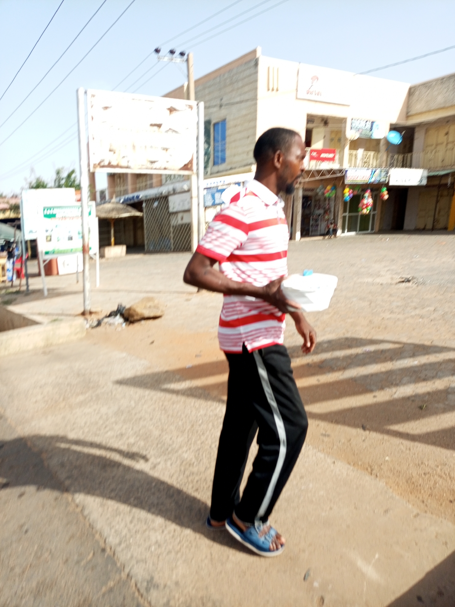 A man is walking on a street in Gombe, Nigeria, holding a white rectangular container. He is wearing a red and white striped shirt, black pants with a white stripe down the side, and blue sandals. The street appears to be paved and sunlit. A building with an Airtel logo is visible in the background, along with various other commercial establishments. There is a sign and some kind of covered structure nearby on the left side of the image. The overall scene suggests an urban, daytime setting.
