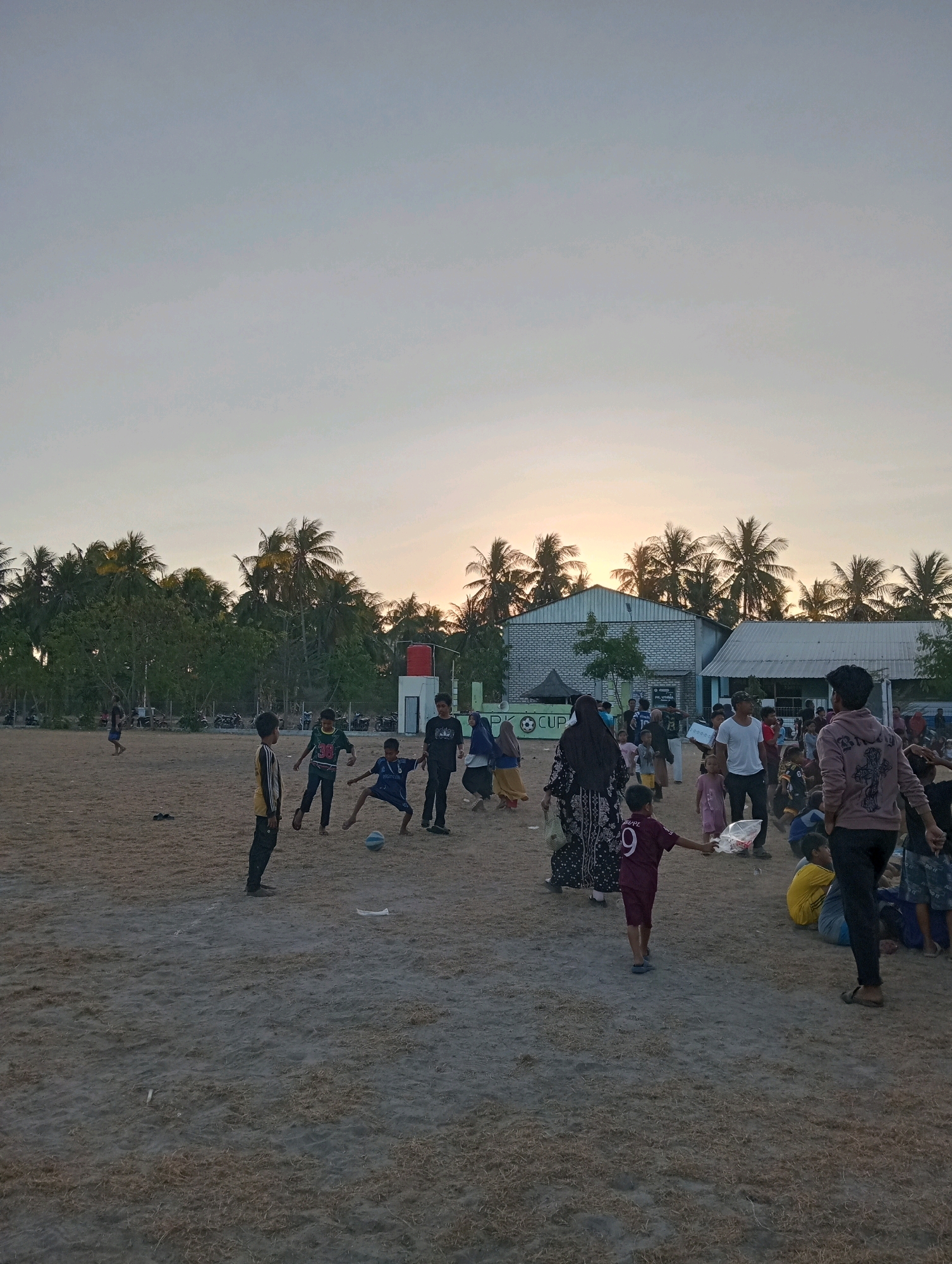 This is a photograph taken in Pagerungan Kecil, Indonesia, showing an outdoor gathering of people on a sandy field, likely a sports ground, at dusk. The sky is a pale gradient of blue and yellow, indicating either sunrise or sunset. Coconut trees form a dense line along the background, behind a few low-rise buildings, one of which has a red water tank on its roof.
Several individuals, predominantly children, are engaged in a game of soccer. One boy in a dark blue jersey and shorts is kicking a soccer ball, while others, including those in yellow, dark green, and black shirts, are positioned around him. Further into the scene, a larger group of people, including adults and children, are milling about. Some are standing, some are sitting, and others appear to be walking. A few individuals are carrying bags. The ground is dry and covered with sparse, dry grass and sand. In the mid-ground, several motorcycles are parked. The overall atmosphere suggests a community event or casual recreational activity.
