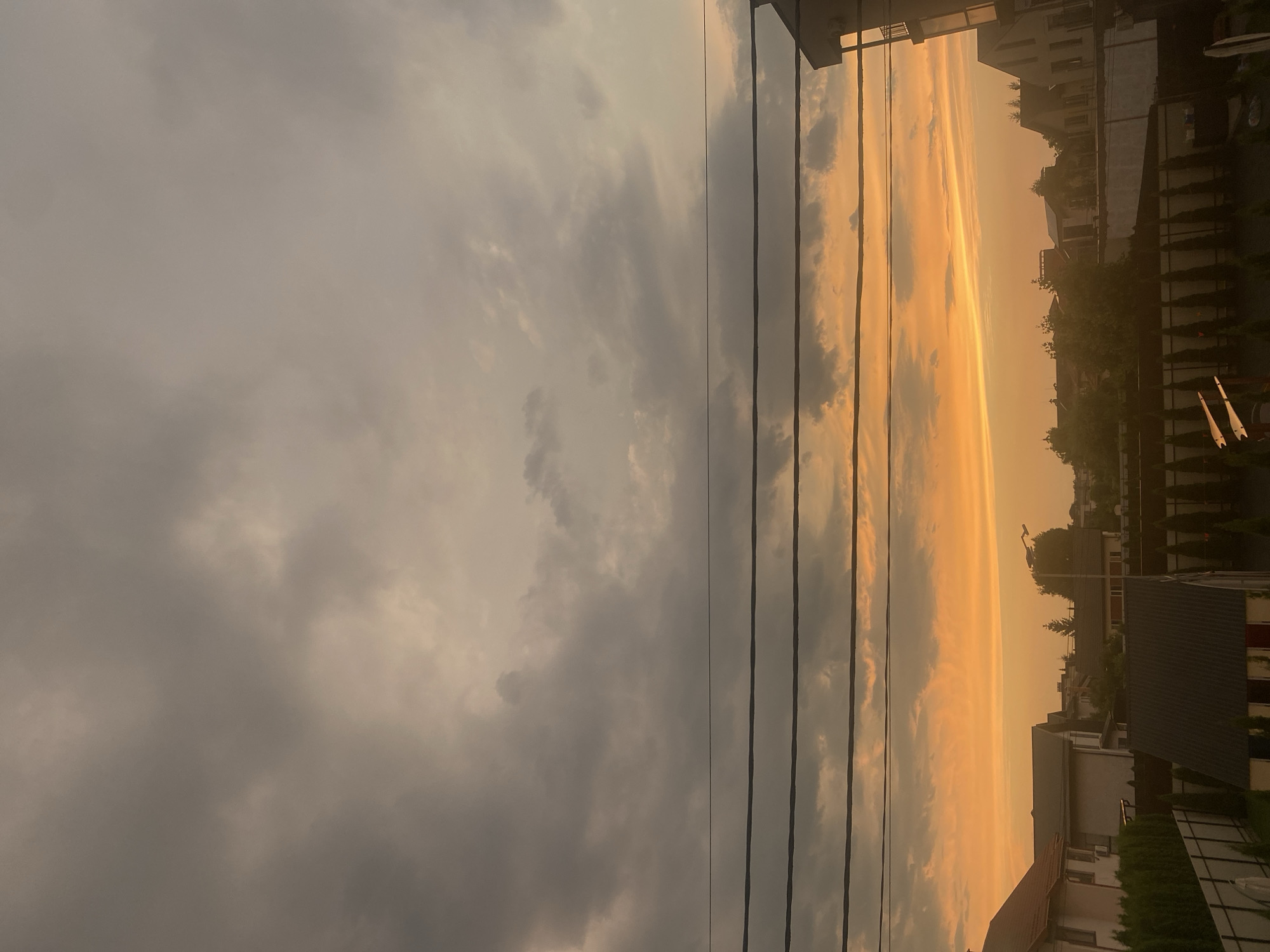 The image depicts a suburban street scene in Novi Petrivtsi, Ukraine, during a sunset. The sky is filled with dramatic clouds, ranging in color from dark gray at the top to a vibrant orange and yellow horizon. Three thick black wires stretch horizontally across the frame, obscuring parts of the sky. Below the wires, a collection of houses with pitched roofs, fences, and lush green trees are visible. The architecture suggests a residential area with varying roof styles and building materials. The lighting indicates the late hour of the day, casting a warm glow over the scene. There are no people or vehicles present in the image.