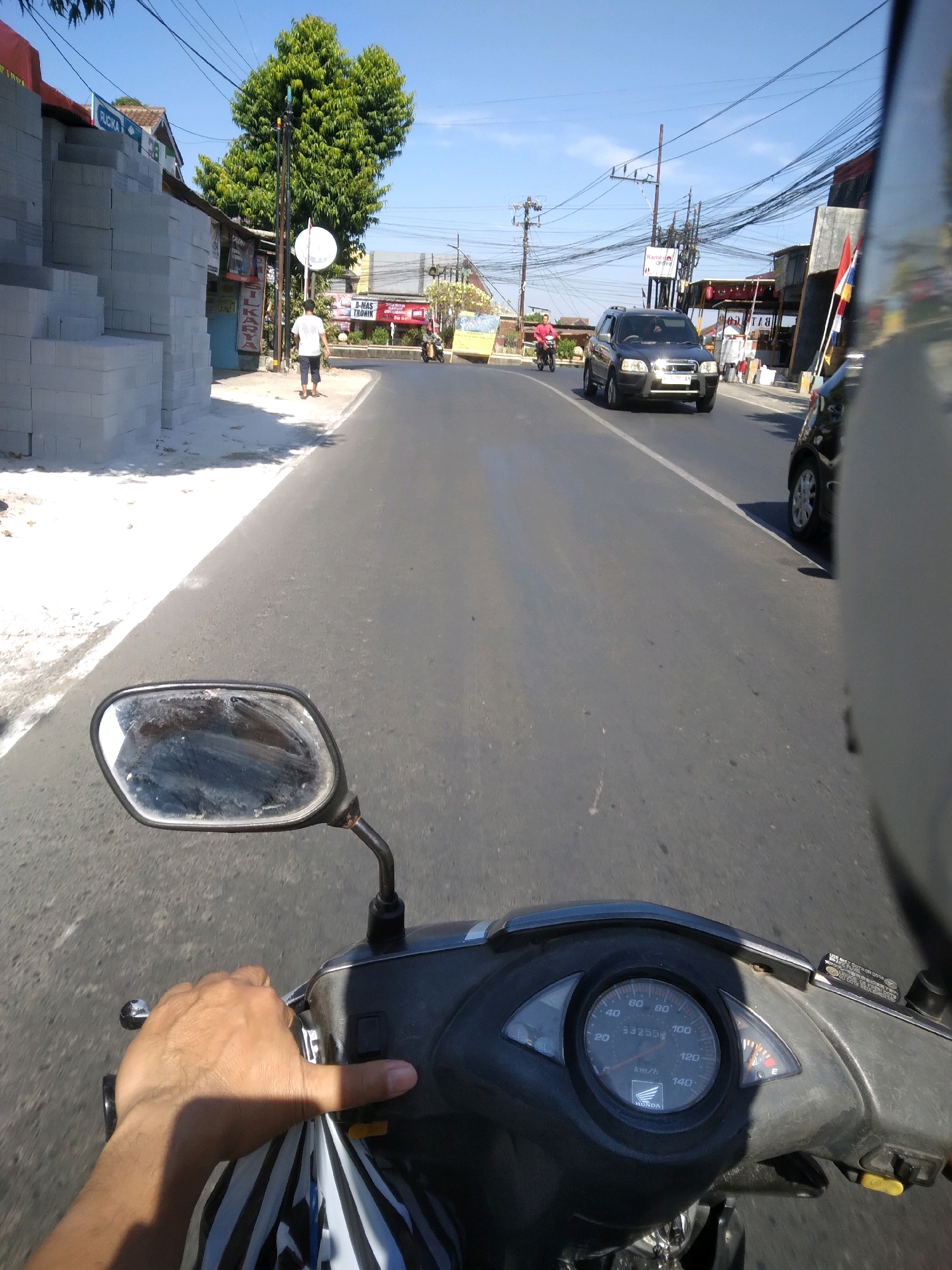 The image depicts a first-person perspective from a motorcycle traveling on a paved road in Purwokerto, Indonesia. The road is lined with buildings and vegetation. To the left, a stack of light gray concrete blocks is visible next to a building with a red sign and a blue door. A white-shirted pedestrian walks away from the camera on the sidewalk. Further down the road, a black SUV is driving towards the viewer, followed by a black car. In the background, a large green tree stands tall against a clear blue sky, with numerous overhead power lines crisscrossing the scene. The motorcycle's dashboard, including the speedometer and fuel gauge, is also visible. The weather appears to be sunny.