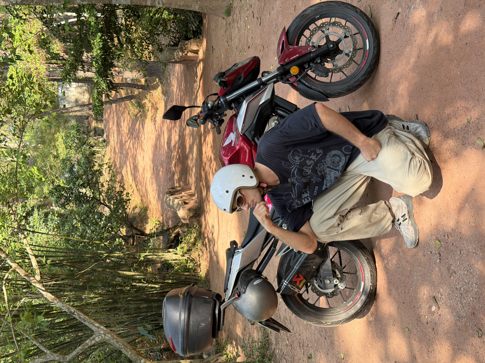 The image shows a man in Thủy Xuân Ward, Vietnam, posed with a red Honda motorcycle. The man wears a white helmet, sunglasses, a dark t-shirt, and khaki pants, and gray shoes. He is leaning on the motorcycle, which is parked on a dirt road or path. The motorcycle has a black storage box mounted on the back. The environment includes trees, bamboo, and a dirt path. The overall setting appears to be a wooded or semi-rural area.