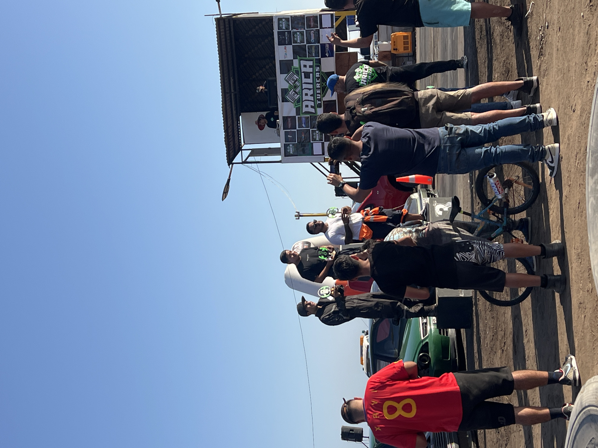A daytime outdoor scene captures a group of people at what appears to be a drag racing event in Arica, Chile. The sky is a clear blue. The finish line structure, prominently displaying the text "ARICA FUERZAS", stands with several spectators gathered around it. The crowd, dressed in casual attire, is observing something beyond the frame of the photo, possibly a race. A bicycle is parked near the spectators. Foreground includes parked vehicles and individuals, one wearing a red shirt with the number "8" on the back. The ground is dirt.