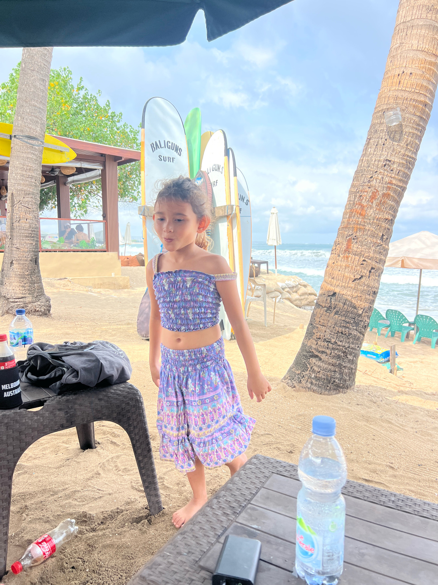 A young girl, appearing to be of East Asian descent, stands on a sandy beach in Kuta, Indonesia, next to a large palm tree. She is wearing a purple, off-the-shoulder, smocked crop top and a matching purple, floral print skirt. Her hair is dark and curly, pulled back from her face.
In the foreground, a dark brown wicker table holds a bottle of water with a blue cap, a black power bank, and a black t-shirt folded on top. To the left of the table, on the sand, lies an empty red and white Coca-Cola bottle. A black cloth bag with "MELBOURNE AUSTRALIA" printed on it is also on the table.
Behind the girl, several surfboards with "BALIGUNS SURF" written on them are propped upright. Further back, beach chairs and umbrellas are visible, along with sandbags stacked near the water's edge. The ocean, with visible waves, stretches out to the horizon under a hazy, partly cloudy sky. A rustic structure, possibly a beachside cafe or bar, is located to the left of the frame, with trees and foliage visible behind it.
