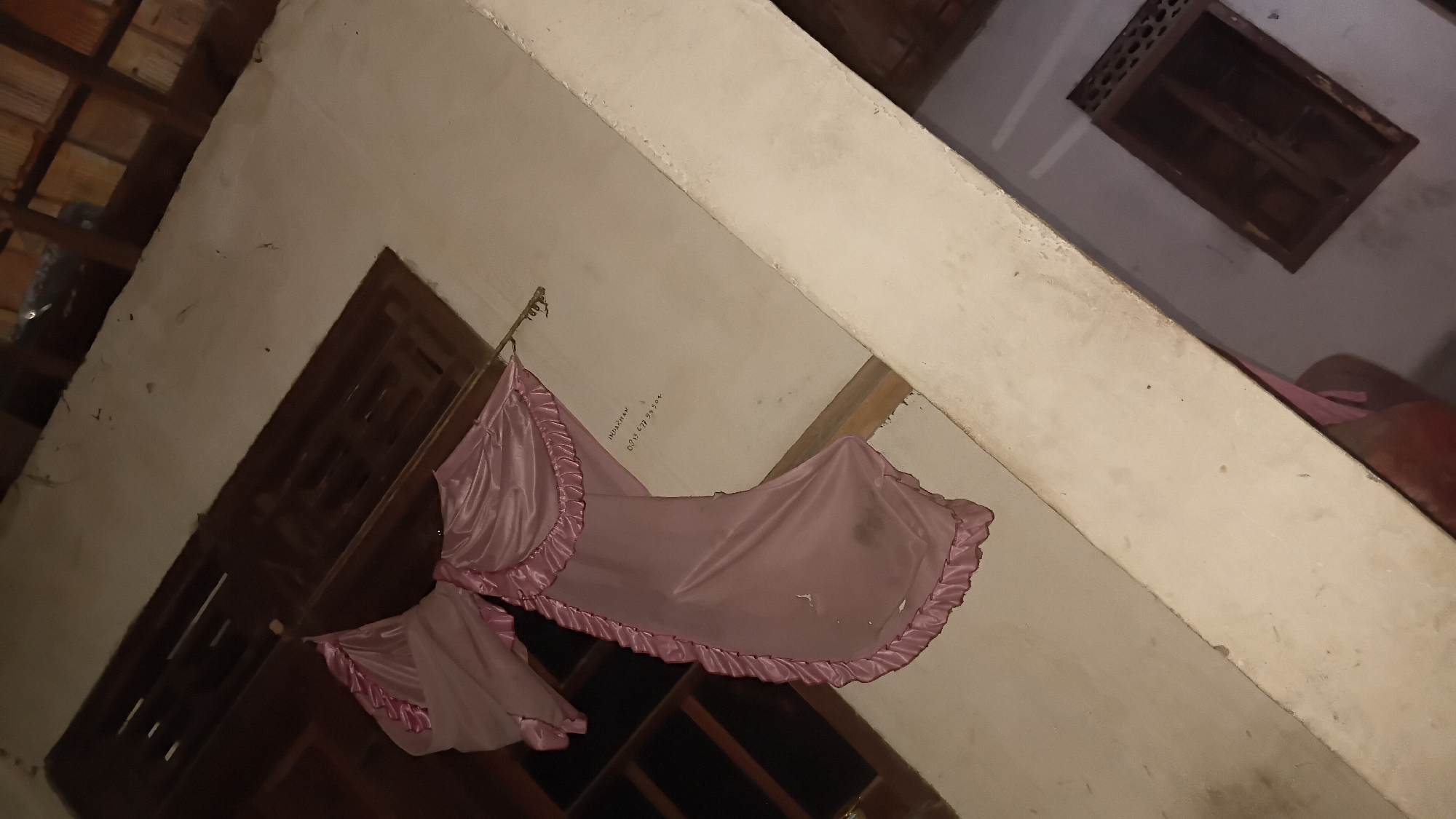 A dusty, pale pink fabric, possibly a curtain or drape, is draped over a metal rod suspended against a cream-colored wall. The fabric has a ruffled edge and is slightly torn in one area. To the left of the fabric, a dark wooden window frame with horizontal slats is visible. Text is scrawled on the wall behind the fabric. The scene appears to be outdoors, possibly a balcony or the side of a building. The overall lighting is dim, suggesting it is either nighttime or heavily shaded. The location is identified as Prabumulih, Indonesia.