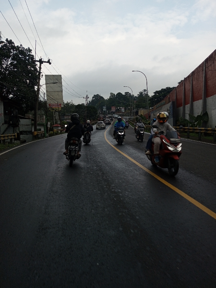 The image depicts a two-lane asphalt road in Dawathang_Dorjibi_Kashingtsawa, Bhutan, under an overcast sky, suggesting recent rain as the road surface is visibly wet. A solid yellow line divides the road longitudinally.
In the foreground and midground, a total of at least nine individuals on motorcycles or scooters are visible. From the low-angle perspective, a dark-clad rider on a dark motorcycle with a visible license plate is positioned in the left lane, moving away from the viewer. Behind this motorcycle, another rider on a similar vehicle follows. In the opposing lane, closer to the viewer, a person on a red scooter wears a light-colored top, light trousers, a helmet with a gold-tinted visor, and blue footwear, appearing to navigate towards the camera. Behind this red scooter, at least five additional motorcycles/scooters with riders, some wearing helmets and rain gear (e.g., a blue raincoat), are also traveling towards the viewer. Further in the distance, more vehicles, including several cars, are present on the road.
The left side of the road features a dark metal guardrail, beyond which dense green foliage and trees are visible. A utility pole with multiple overhead power lines stands prominently. Behind the pole, a tall, light-colored rectangular billboard or structure is present, with a smaller, partially visible sign below it featuring red text on a white background. Further back, more trees and hints of urban structures are visible.
The right side of the road also has a dark metal guardrail. Beyond this, a large retaining wall with distinct red horizontal bands separated by vertical grey or white pillars extends into the distance. A streetlight curves over the road from this side. The sky is uniformly grey and cloudy.