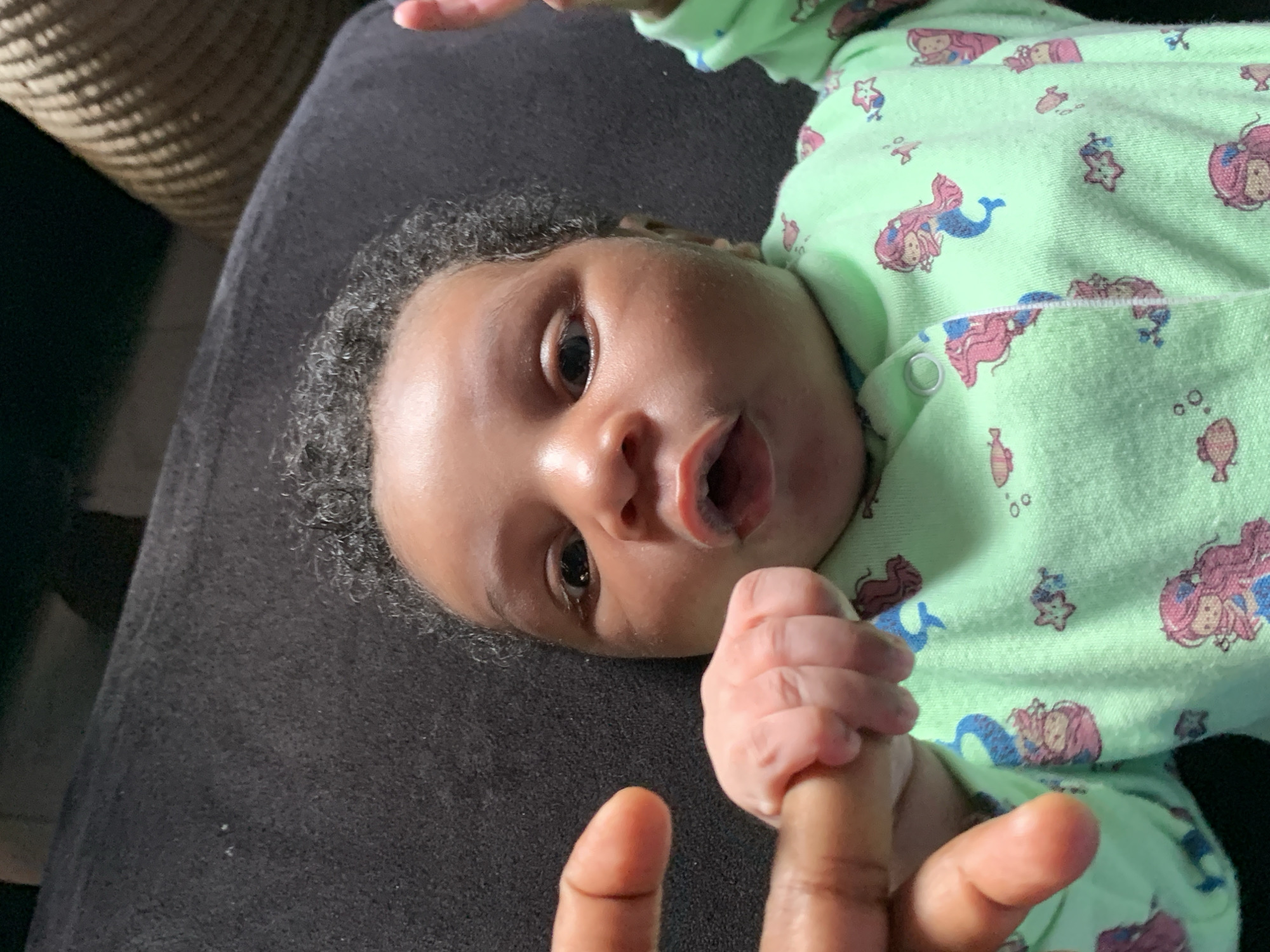 A close-up, slightly tilted image captures an infant lying on a dark grey or black textured surface, likely a blanket or furniture. The baby has dark, curly hair and dark skin, with wide, dark eyes looking directly at the viewer. The infant's mouth is slightly open, hinting at an expression of curiosity or an attempted sound.
The baby is dressed in a light green pajama or romper covered with whimsical patterns of mermaids, starfish, and small fish, primarily in pink, blue, and brown tones. The garment has visible snap buttons along the side.
In the foreground, the baby's small hand is firmly gripping an adult's index finger, showcasing a tender moment of connection and the infant's natural grasping reflex. Only a portion of the adult's hand is visible. The background, particularly in the upper left, features a blurred, textured object, possibly a woven basket or furniture piece, in a warm, earthy tone. The setting appears to be indoors, under ambient lighting. The scene conveys a sense of warmth and interaction between the infant and caregiver. No visible text is present. This image was taken in Ibasa, Nigeria.