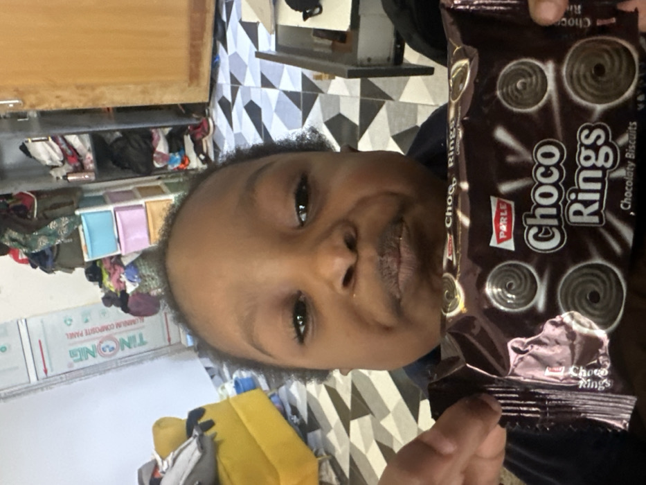 A young individual with dark complexion and short, dark hair, wearing a dark garment, is depicted in a medium shot, looking directly towards the camera with a slight pout. They are holding a brown, foil-wrapped package of "Parle Choco Rings" Chocolaty Biscuits in both hands, positioned below their chin. The background reveals an indoor residential setting in Ilishan, Nigeria. The floor features black and white geometric patterned tiles. To the left, a yellow upholstered armchair is partially visible. Further back and to the right, a multi-tiered plastic drawer unit, containing various items, stands next to a wooden door. A white wall section to the left displays a red and white sign for "TINONG ALUMINIUM COMPOSITE PANEL." The environment appears lived-in.