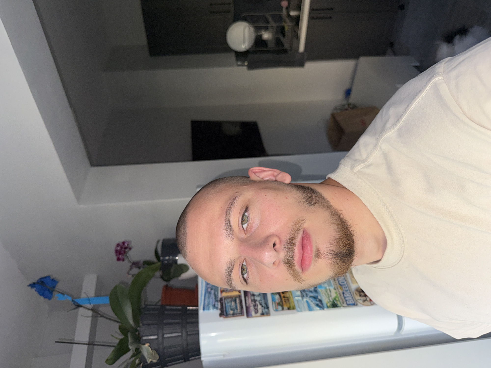 A man with a shaved head and a goatee is posing for a selfie indoors. He is wearing a light-colored t-shirt. Behind him, to his left, is a white refrigerator adorned with magnets, and above that, a shelf holding potted plants, one with a blue orchid. To his right, a doorway leads to a dimly lit area with kitchen cabinets and appliances. The scene appears to be in a residential setting, potentially in the city of Stara Zagora, Bulgaria.