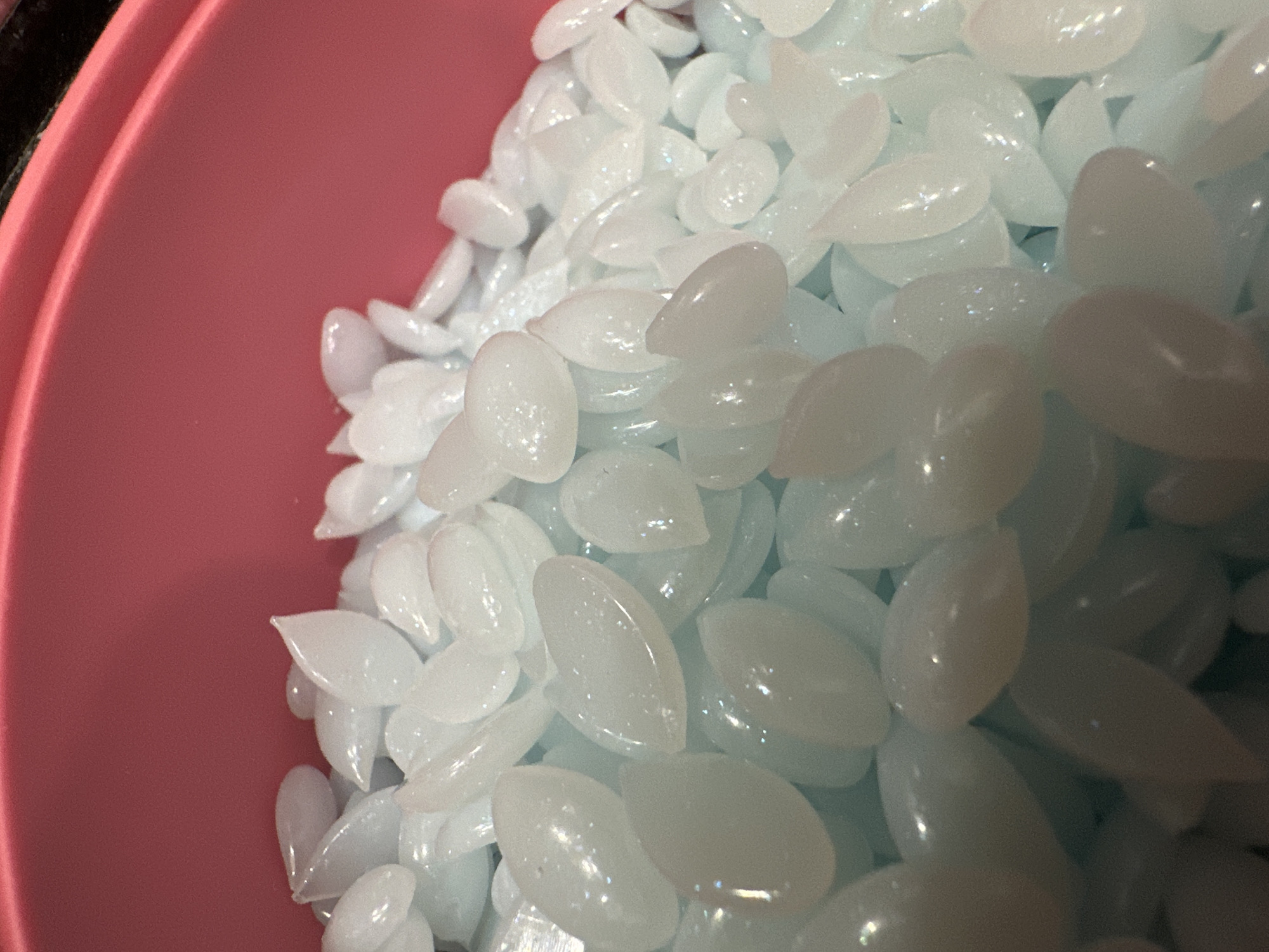 The image shows a close-up view of a pile of white, opaque, oval-shaped beads, possibly polymer pellets or decorative stones. They are contained within a pink plastic container, the rim of which is visible at the top and right edges of the frame. The beads have a slightly iridescent quality, catching the light. There are no people, actions, or interactions depicted. The setting is an indoor environment, with no discernible urban features of Shchyolkovo, Russia.