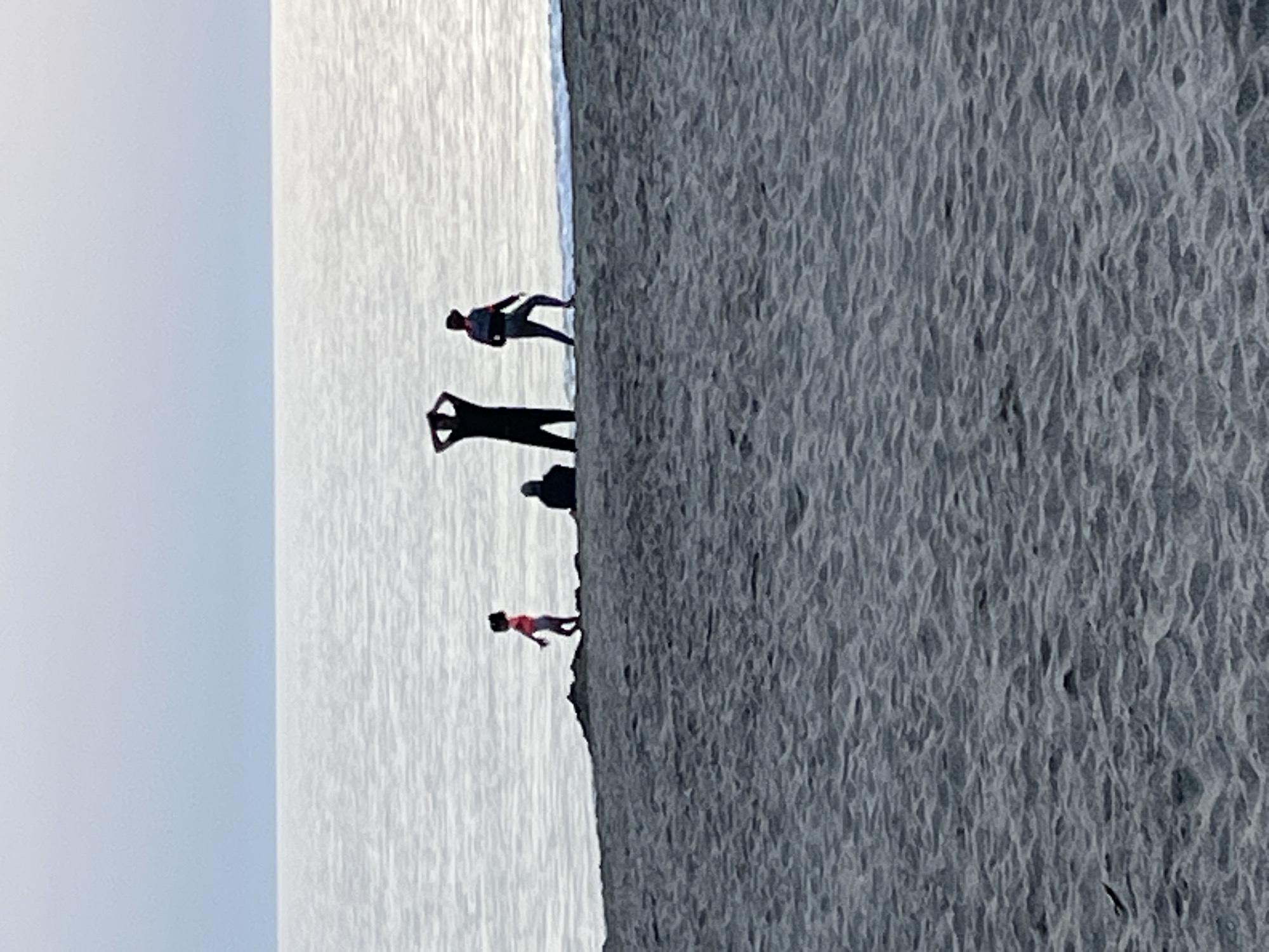 The image depicts a sandy beach in Alanya, Turkey, with the ocean on the left side of the frame. Four individuals are visible along the shoreline. The person closest to the ocean is facing forward, and has their hands on their head. The second person is walking. The third person is a shadow, and the last person is further down the beach, walking along the shore. The sand appears textured with patterns or ripples. The sky is overcast.