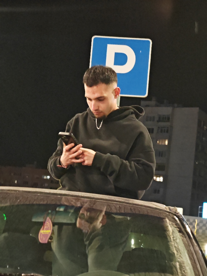 The image depicts a nighttime scene in an urban environment in Shumen, Bulgaria. A young adult male is centrally positioned, observed from the chest up, with his lower body obscured by the foreground. He has short, dark hair with faded sides and is wearing a dark gray or black hooded sweatshirt over a silver-colored chain necklace. A red string bracelet is visible on his left wrist. He is actively engaged with a dark-colored smartphone, holding it vertically with both hands and looking downwards at its screen.
In the immediate foreground, the top section of a vehicle, specifically the windshield and roofline, partially obstructs the view of the male subject. The windshield appears to have a layer of dust or dirt. A purple, leaf-shaped car air freshener with yellow text is suspended inside the vehicle, visible through the windshield, and a partial reflection of the male subject can be seen on the glass.
Behind the individual, a blue square parking sign with a white capital "P" is mounted on a pole against a dark background. Further in the distance, multi-story residential buildings with several illuminated windows are visible under the dark sky. The precise street or district within Shumen, Bulgaria, is not discernible from the visual evidence provided.