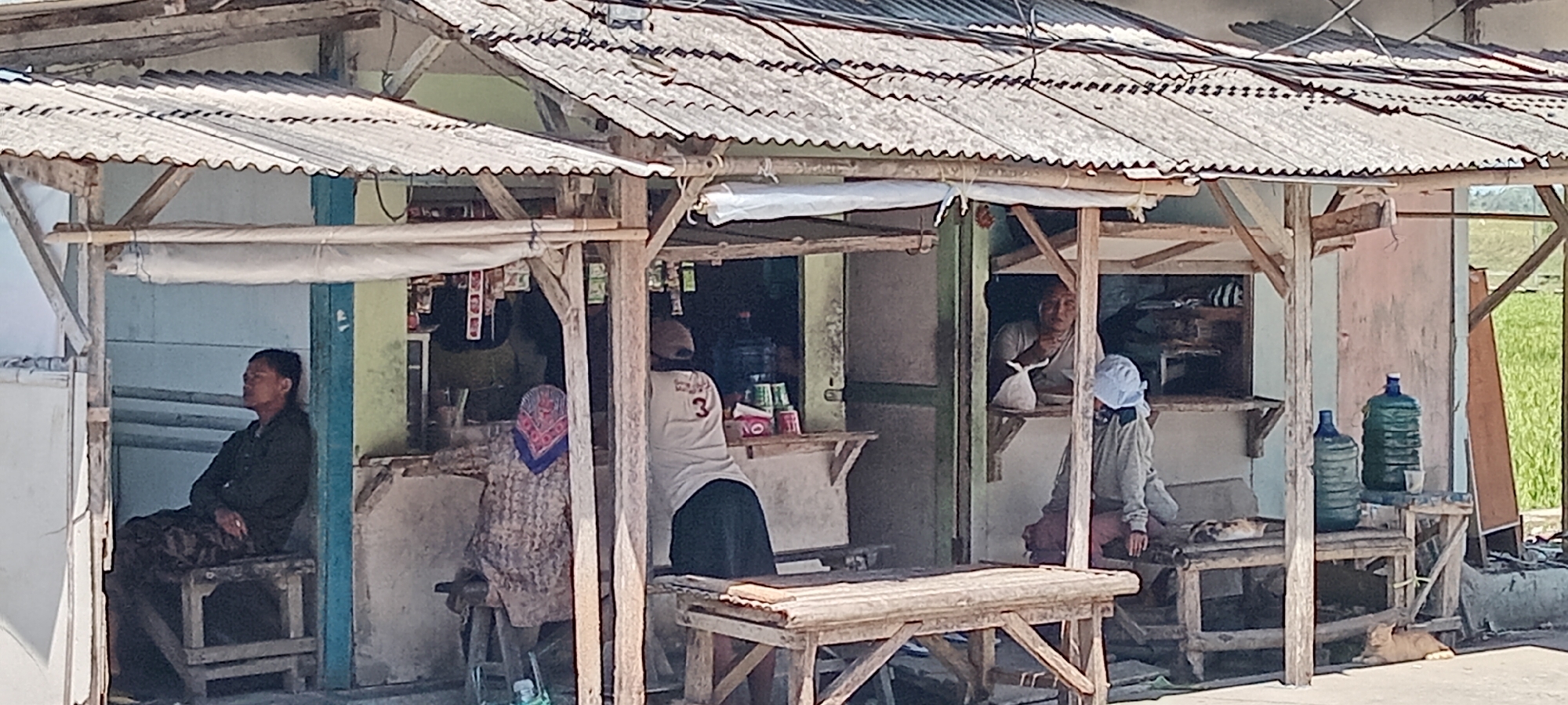 A rustic, open-air shop in Maruyung Sari, Indonesia, is depicted. The structure features a corrugated metal roof supported by wooden beams, with two distinct stalls. In the left stall, a man is seated on a wooden stool, facing left with his eyes closed. He wears a dark long-sleeved shirt and patterned trousers. In the center stall, a woman wearing a patterned headscarf and clothing is seated, facing away from the camera. Behind her, a person in a light-colored shirt with the number "3" on it and a dark skirt stands, facing the camera. To the right, an elderly person with a light-colored head covering is visible behind a counter, holding a white plastic bag. Another individual, also wearing a light-colored head covering and a light-colored jacket, is seated in the right stall. Three large plastic water bottles, two blue and one green, are arranged on a wooden shelf to the right of the right stall. Two orange cats are visible in the foreground; one is lying on a wooden surface in the right stall, and another is lying on the ground in front of the shop. The shop appears to be selling various goods, as indicated by items displayed on shelves within the stalls. The background shows a green, possibly agricultural, area.