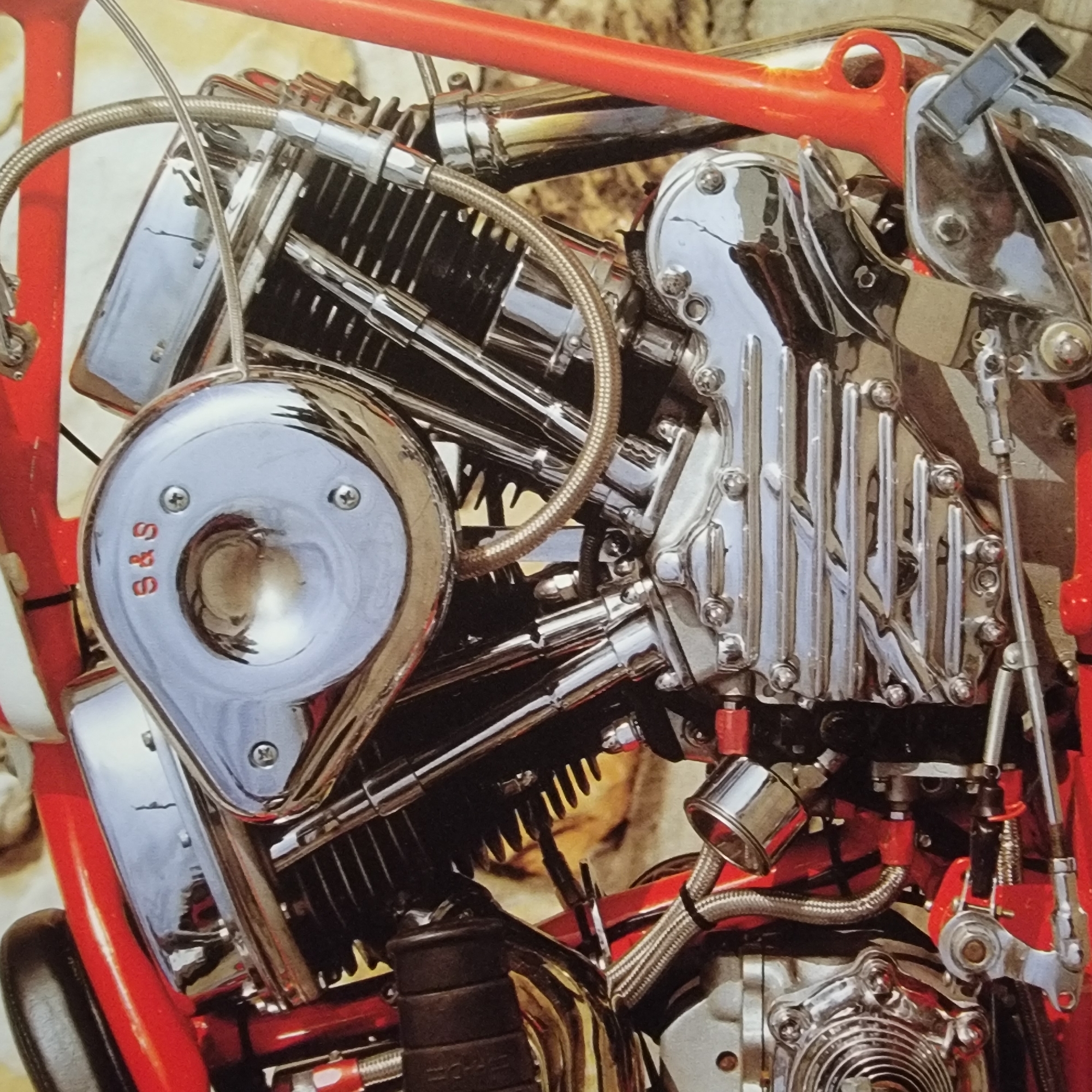 The image is a close-up, eye-level, medium shot of the engine of a custom motorcycle. The motorcycle is predominantly red and chrome. The engine is a V-twin, with prominent chrome details. The air cleaner cover is circular and chrome, with the letters "S & S" in red. There are also various hoses, wires, and mechanical components visible, all rendered in high detail with a focus on reflective surfaces. The background is blurred and appears to be textured, possibly a stone or concrete surface, with some yellowish-brown tones. No people or identifiable landmarks are present, and the location is unknown.