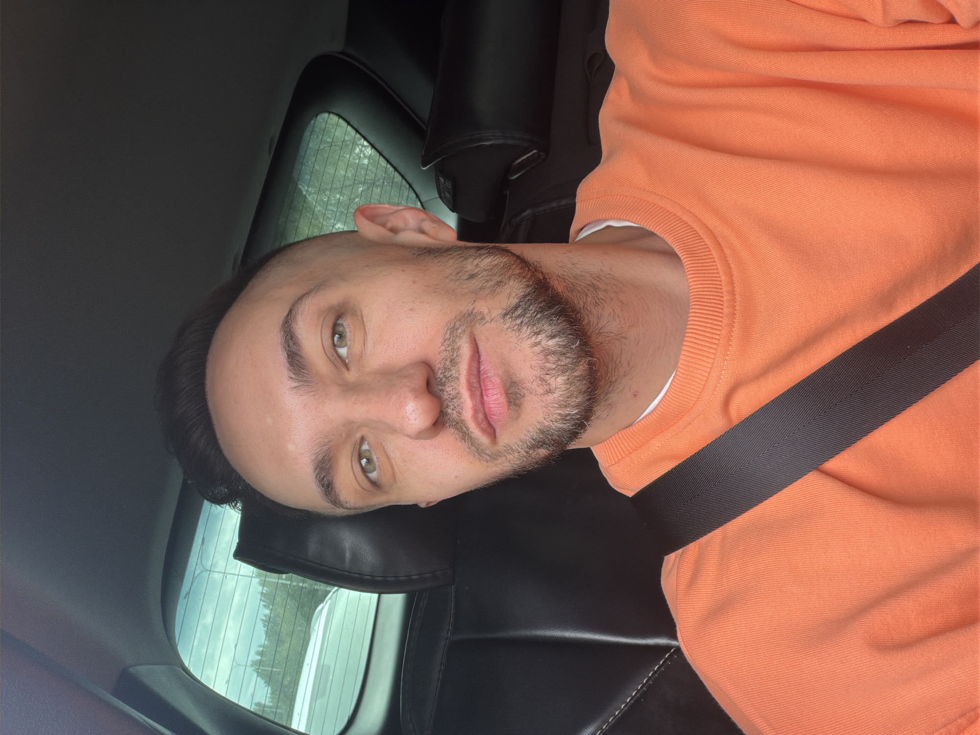 The image is a close-up, selfie-style shot of a man seated in the passenger seat of a car. He has light skin, light green eyes, a short haircut with shaved sides, and a beard. He is wearing an orange sweatshirt and a black seatbelt is visible across his chest. The background shows the interior of a car, including black leather seats and the rear window, through which trees and a cloudy sky are visible. The exact location within Astana, Kazakhstan is not discernible from the image.