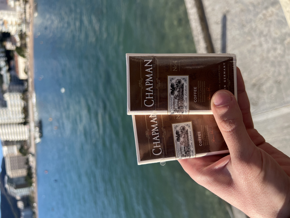 A hand holds two packs of "Chapman Coffee" cigarettes against a backdrop of a body of water and a distant shoreline. The cigarette packs are brown and white with text stating "Chapman No. 4" and "With von Eicken". Further text mentions "Hand selected tobaccos blended to perfection" and "roasted coffee aroma". The bottom of the packs state "COFFEE" and "Premium Cigarettes Made in Germany". The shoreline appears to show buildings and trees. The water surface has ripples.