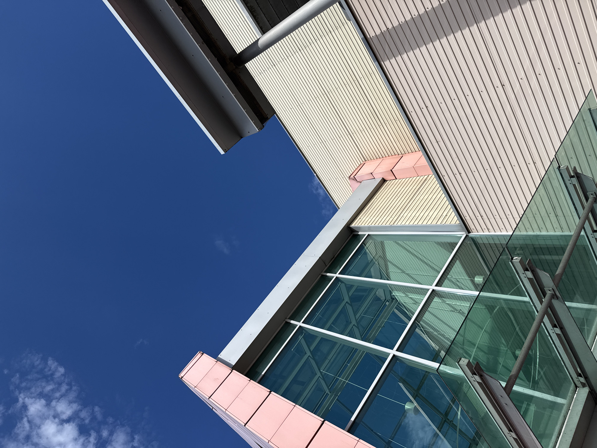 This is a low-angle, full-frame, eye-level, outdoor, daytime, natural light, color photograph of a modern building's exterior, with a deep blue sky and sparse white clouds in the background. The building's architecture is characterized by sharp angles and contrasting materials. A large section of the building features a glass facade with a silver-colored metal frame. Above and to the right of the glass section, there is a cream-colored corrugated metal wall, and further to the right, a silver-colored overhang supported by a cylindrical metal pillar. A pink-colored accent piece is visible on the corner where the glass and cream-colored wall meet. The photograph does not depict any people or activities. The location is stated to be Bielany Wrocławskie, Poland.