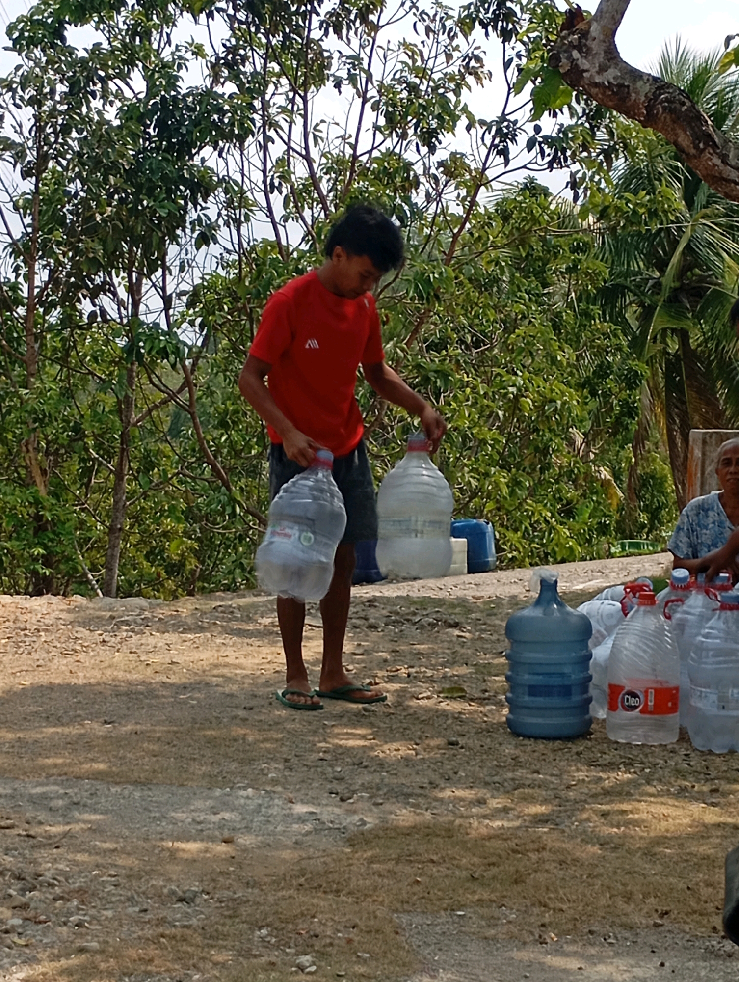 In Ketepung, Indonesia, a man in a red t-shirt and dark shorts is carrying a large, clear plastic water jug, its contents sloshing. He is barefoot, wearing flip-flops on his feet. To his right, several more water jugs are visible, some of them clear and others blue. A woman, wearing a blue and white patterned top, sits in the background, facing the jugs. The scene appears to be outdoors, with lush green trees and vegetation surrounding the area, and the ground is covered in dirt and gravel. The lighting suggests it is daytime, with shadows cast on the ground.