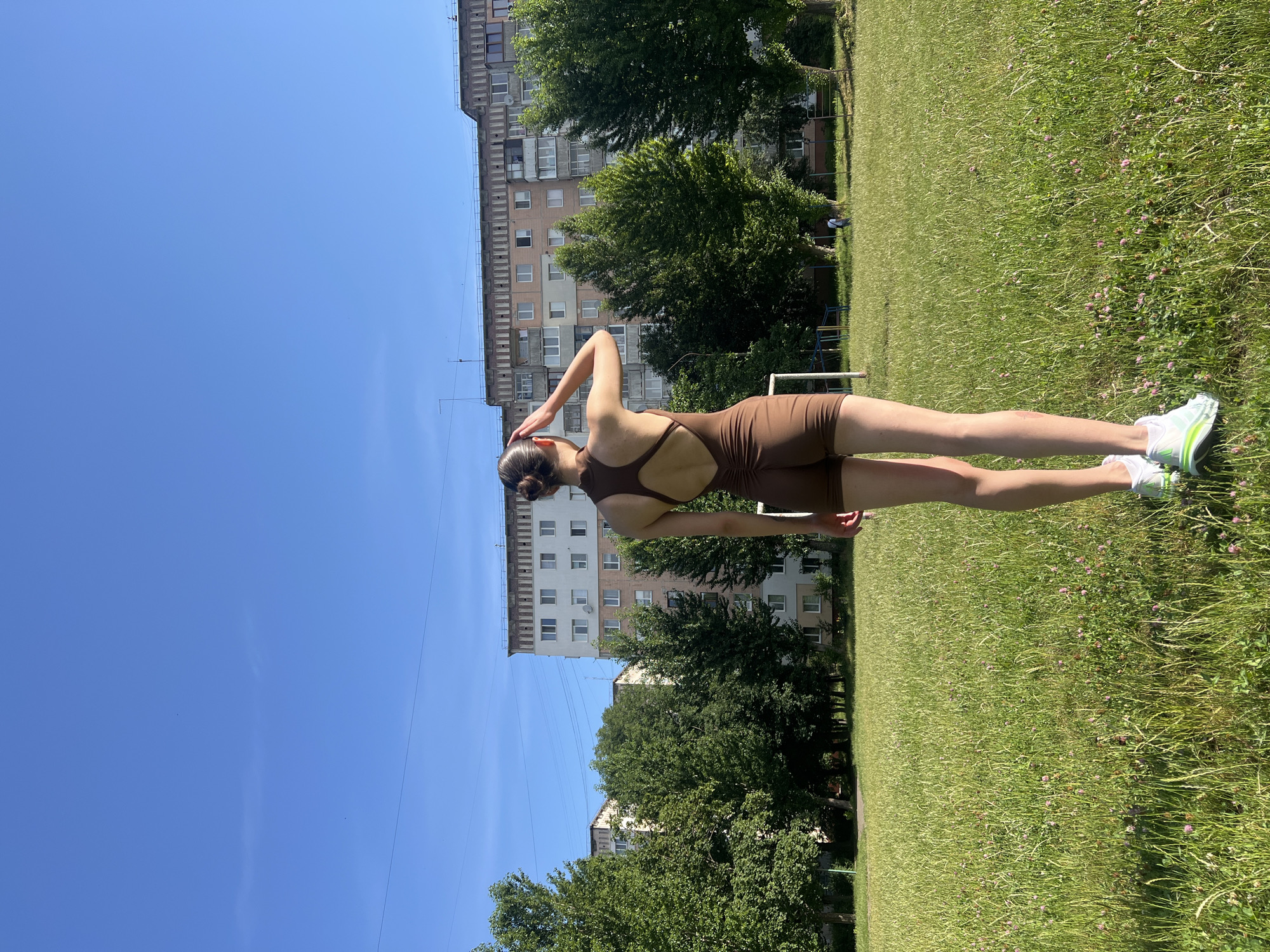 A young woman with her hair in a bun is standing in a grassy field with a building and trees in the background. She is wearing a brown one-piece athletic outfit and white sneakers with green accents. The woman is facing away from the camera, with her right arm raised and her hand on the back of her head. Her left arm is bent at the elbow and her hand is resting on her hip. She is looking over her left shoulder. The sun is shining brightly, casting shadows on the ground, suggesting it is daytime. The sky is clear and blue. The building in the background appears to be an apartment building. There are several trees scattered throughout the scene. The grass is green and appears to be well-maintained, with some small pink flowers scattered throughout. This image was taken in Ternopil, Ukraine.