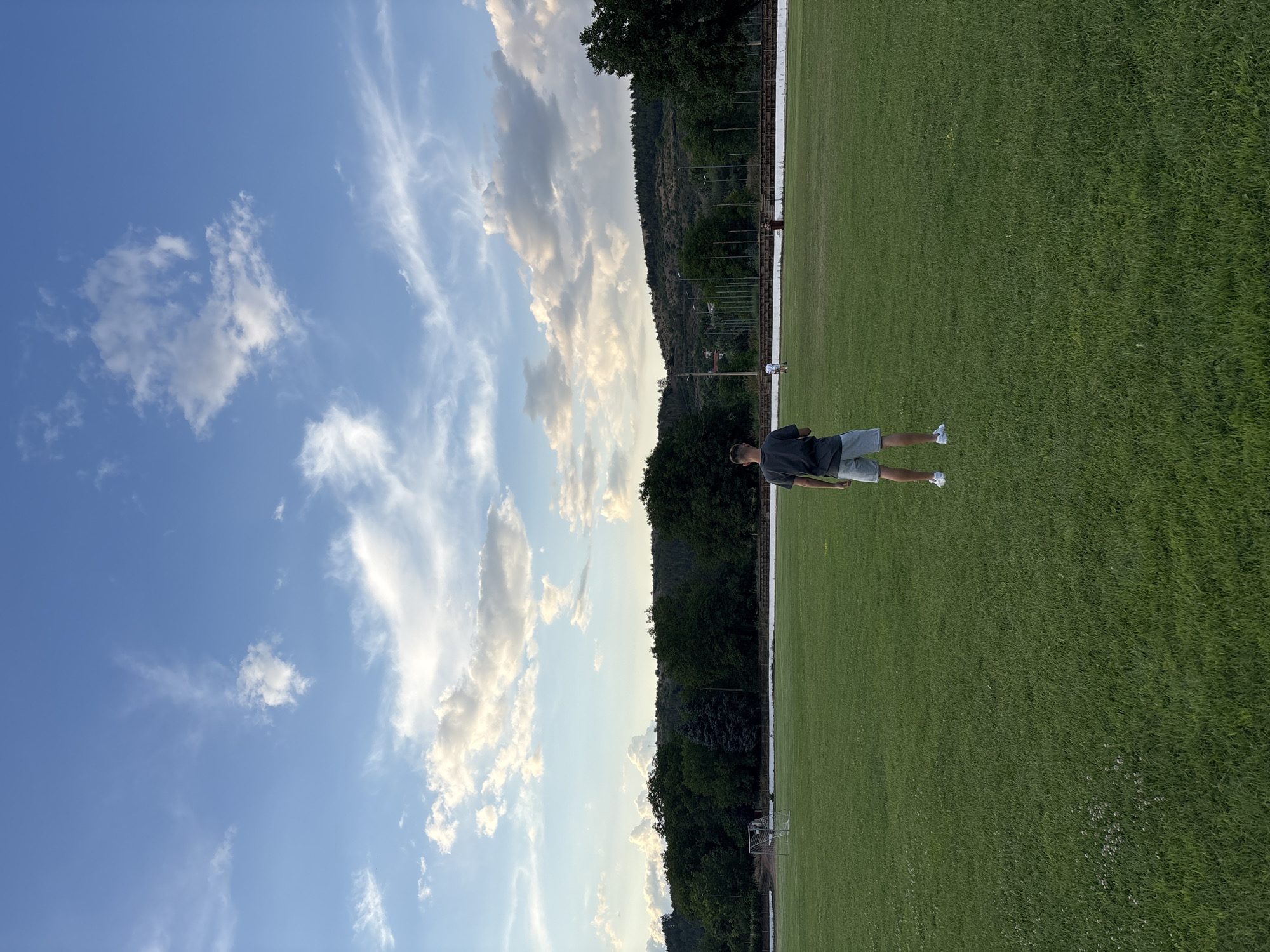 In Boboshevo, Bulgaria, a lone individual stands on a lush green field, facing away from the camera. The person is wearing a dark t-shirt and light-colored shorts, with white sneakers. The field appears to be a sports ground, with a goalpost visible in the distance to the left. Beyond the field, a line of trees forms a dense barrier, with hills covered in vegetation rising behind them. The sky above is a vibrant blue, dotted with scattered white clouds, suggesting fair weather. The overall impression is one of an open, rural outdoor space.