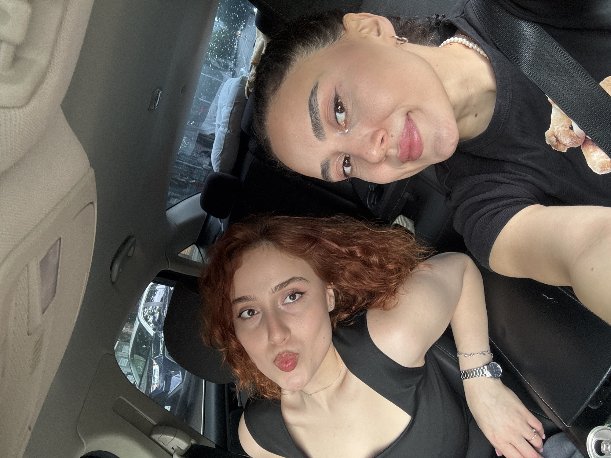 Two women pose for a selfie inside a car. One woman is brunette with her hair pulled back, wearing a black T-shirt with a bear design and a pearl necklace; she has a mole near her lip. The other woman has curly red hair and is wearing a black top with a geometric neckline, a watch, and a thin necklace. Through the car windows, other vehicles and buildings suggest an urban environment, identifiable as Tbilisi, Georgia. Raindrops streak the windows.