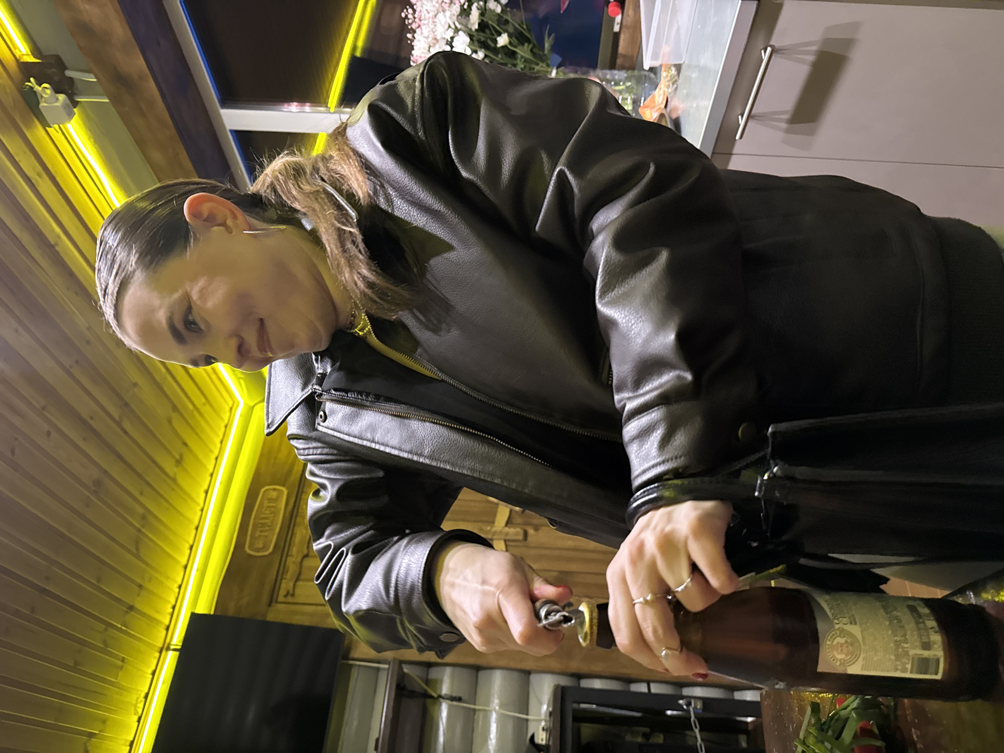A woman in a black leather jacket is opening a brown glass beer bottle with a bottle opener. She is wearing multiple rings on her fingers. The setting appears to be indoors, with a yellow-lit wooden wall visible in the background. Part of a TV screen and cylindrical objects, possibly kegs, are also visible. Based on the photo's context, the location is likely in Perm, Russia.