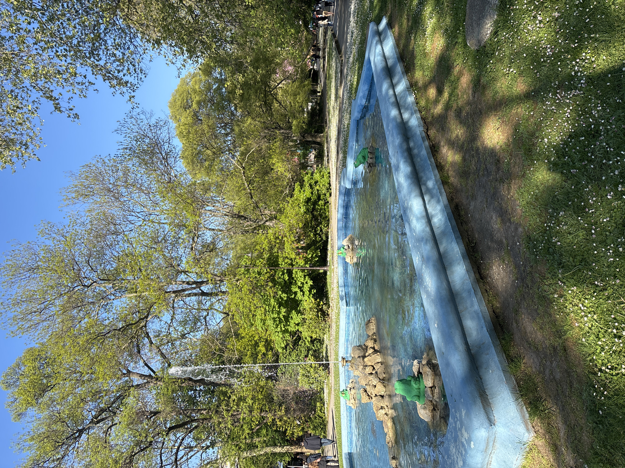 The image shows a long, blue, multi-tiered fountain in a park-like setting. The fountain contains water, and there are green frog statues placed on rock-like features within it. A jet of water is spraying upwards from the fountain. The surrounding area features green grass dotted with small white flowers, trees with green foliage, and a paved walkway. Some people are walking along the walkway in the background. The sky is visible through the trees and is a clear blue.
The fountain is located in the Sea Garden of Varna, Bulgaria, near the children's playground "The Frog". The fountain is a long, rectangular structure with multiple levels, painted a distinctive shade of blue. There are several green frog statues placed on rocks or platforms within the fountain.