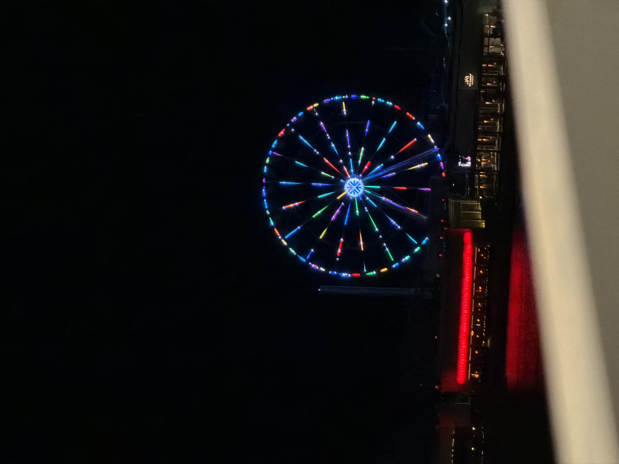 In the city of Unknown, Unknown, at night, a large Ferris wheel with colorful lights stands against a dark sky. In the foreground, a body of water reflects the red lights of a building that stretches across the lower portion of the frame. The building appears to be a restaurant or entertainment venue with several illuminated windows. A railing is visible in the extreme foreground, suggesting the photo was taken from an elevated vantage point. The overall scene depicts a vibrant night-time atmosphere, likely a waterfront amusement area.