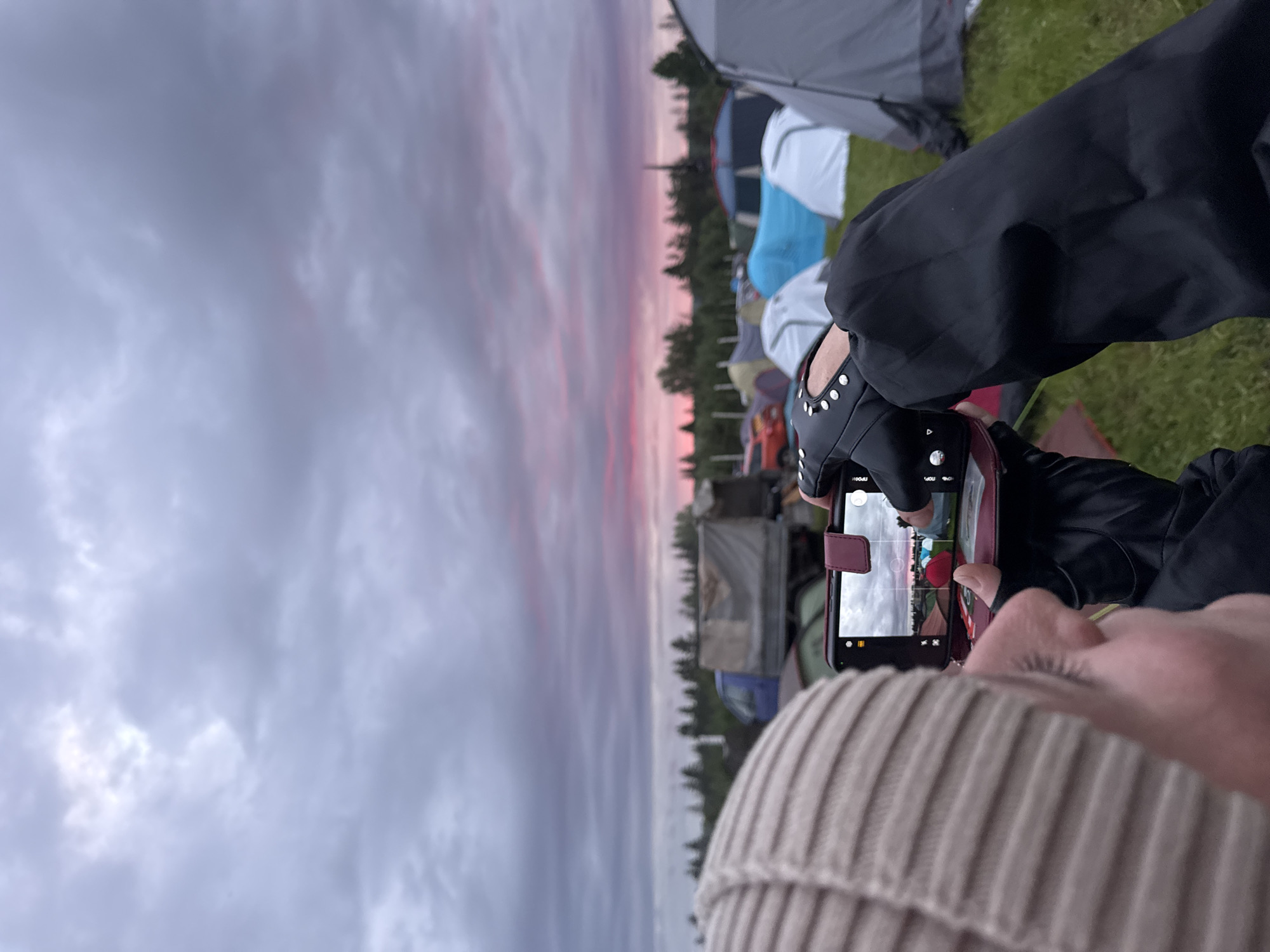 A partial view of a person captures a twilight scene at a camping ground in Большая Кивара, Russia. The person, viewed from behind the right shoulder, wears a light-colored ribbed beanie and a dark, possibly leather, glove with metallic stud detailing on the wrist. They hold a smartphone horizontally, encased in a maroon flip case, displaying an active camera interface. The phone's screen shows a live preview of the scene ahead, mirroring some of the visible elements.
The background reveals a large grassy field interspersed with numerous tents of various colors, including grey, white, blue, and red. Beyond the tents, several vehicles, including trucks with covered beds and what appear to be smaller cars, are visible. A dense line of evergreen trees forms the distant horizon. The sky is overcast with thick clouds, transitioning from lighter grey at the top to dramatic purplish-pink and orange hues near the horizon, indicative of either sunrise or sunset. The overall lighting suggests dusk or dawn conditions.