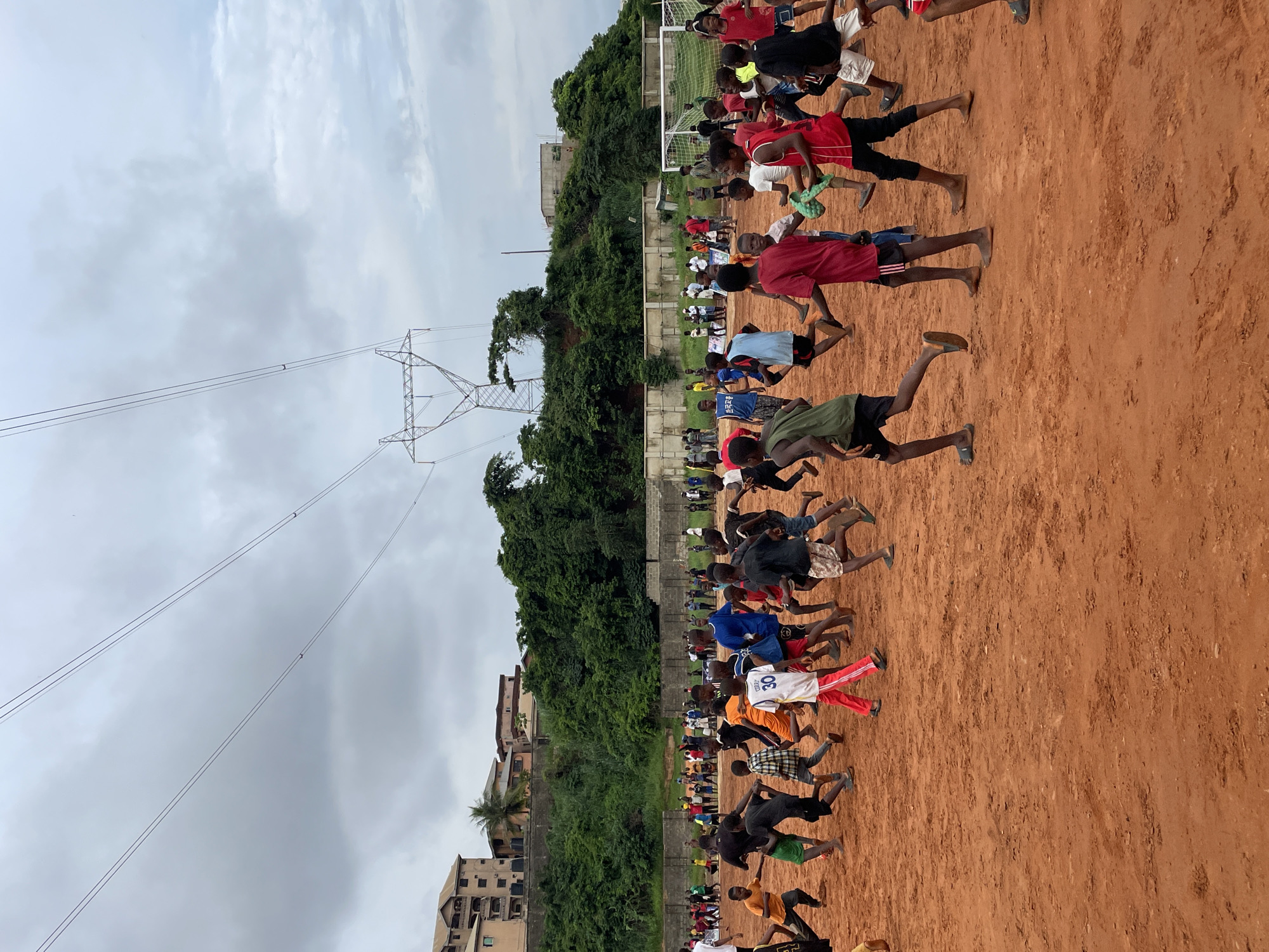 A group of boys are playing soccer on a dirt field. The field is surrounded by a fence, and in the background, there are buildings and trees. The sky is cloudy.
