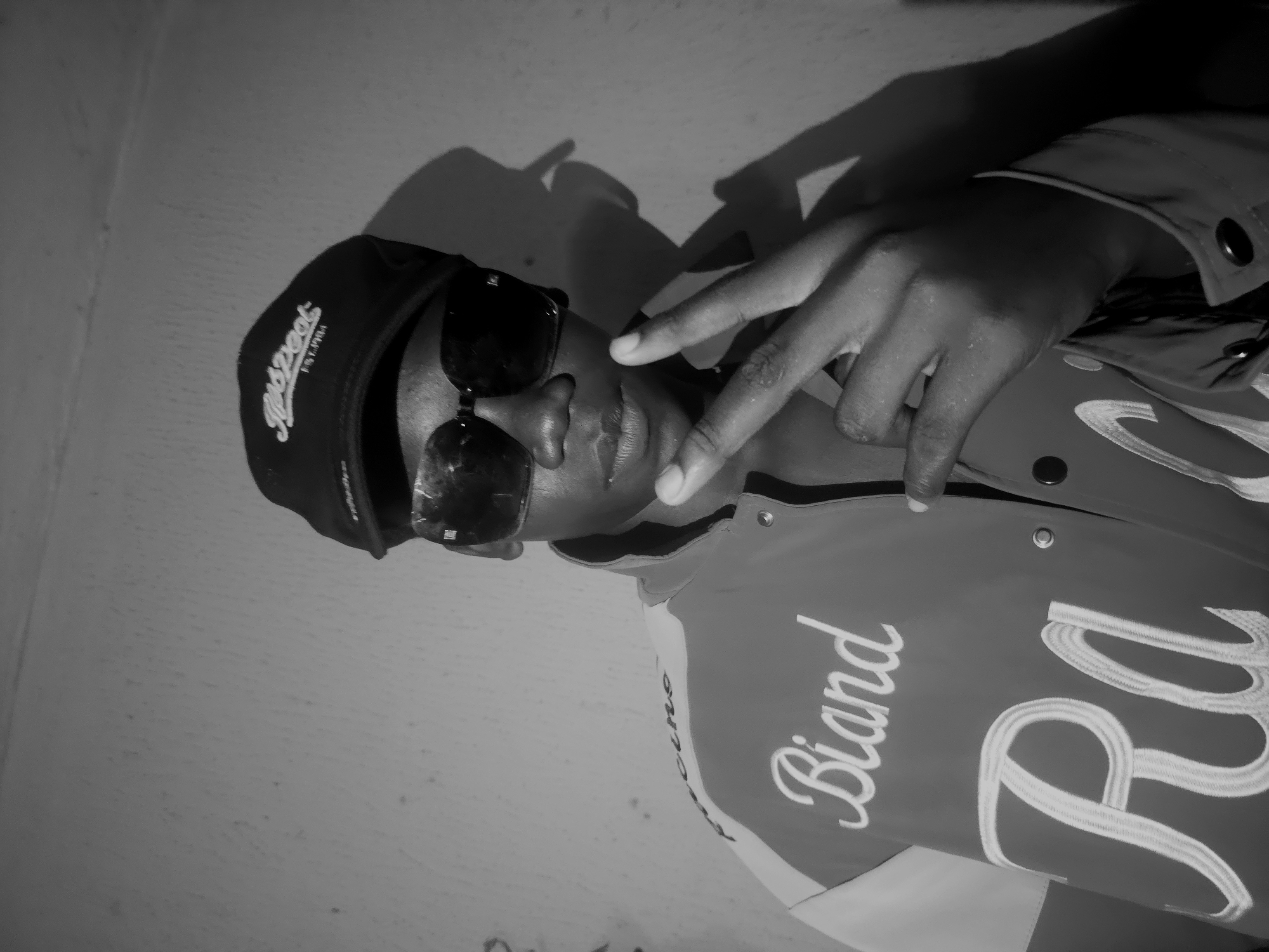 A black and white portrait features a person, likely male, positioned against a plain wall, possibly indoors or in a sheltered outdoor setting. The image is taken at a slight angle, with the subject leaning or posing. The person has dark skin and is wearing a dark baseball cap with the word "Redpeat" stylized in lighter script and "EST.1994" underneath it. Dark, square-framed sunglasses obscure their eyes.
The subject is also wearing a jacket, possibly a varsity or bomber style, with prominent embroidered text. Visible text on the jacket includes "Biand" in a script font on the left chest area, and a large, stylized "Ra" in a similar embroidered style to the right. The jacket appears to have snap buttons down the front. The person's left hand is raised to head level, fingers extended in a V-shape gesture. Distinct shadows are cast on the wall behind the subject, suggesting a direct light source. The overall mood conveyed is one of confidence or coolness. There are no visual cues within the image itself to specify the location as Asaba, Nigeria, but this is the provided context.