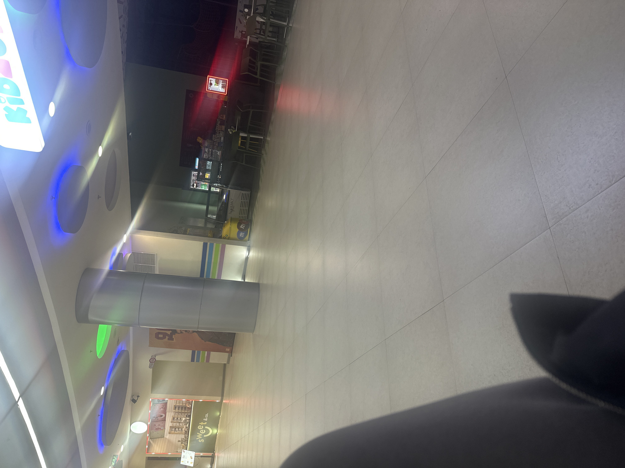 The image shows a brightly lit indoor space, likely a shopping mall, in Yerevan, Armenia. A large cylindrical support column is visible, with a store labeled "sweet & co." nearby. In the distance, there is a food court area with tables and chairs, and a sign with what appears to be a food advertisement. The foreground includes a dark, unidentifiable object, possibly clothing. The floor is tiled in a neutral color.