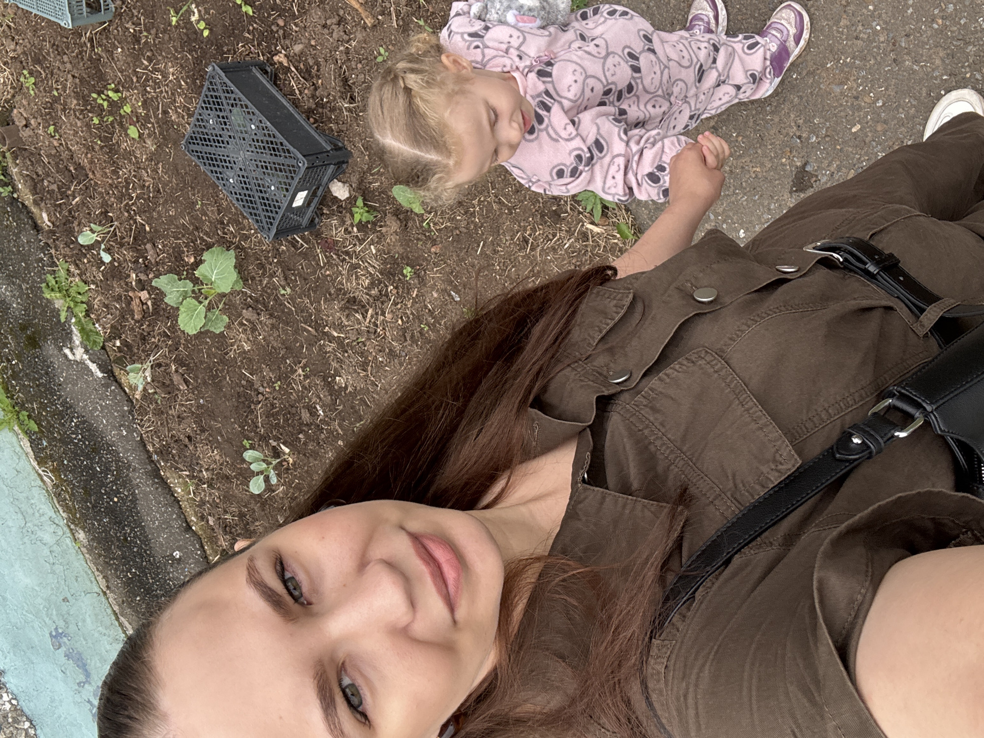In Chaikovsky, Russia, a woman with brown hair pulled back takes a selfie in an outdoor, unpaved area, possibly a garden or allotment. She is wearing a brown button-up garment. Next to her, a young child with blonde hair in braids is holding her hand and wearing a pink pajama suit with a cartoon bear pattern and purple sneakers. The child is looking down. In the background, there is dark soil with sparse green plants, dry twigs, and a black plastic crate. A greenish-blue painted concrete edge runs along the top of the frame. The overall lighting suggests an overcast day.