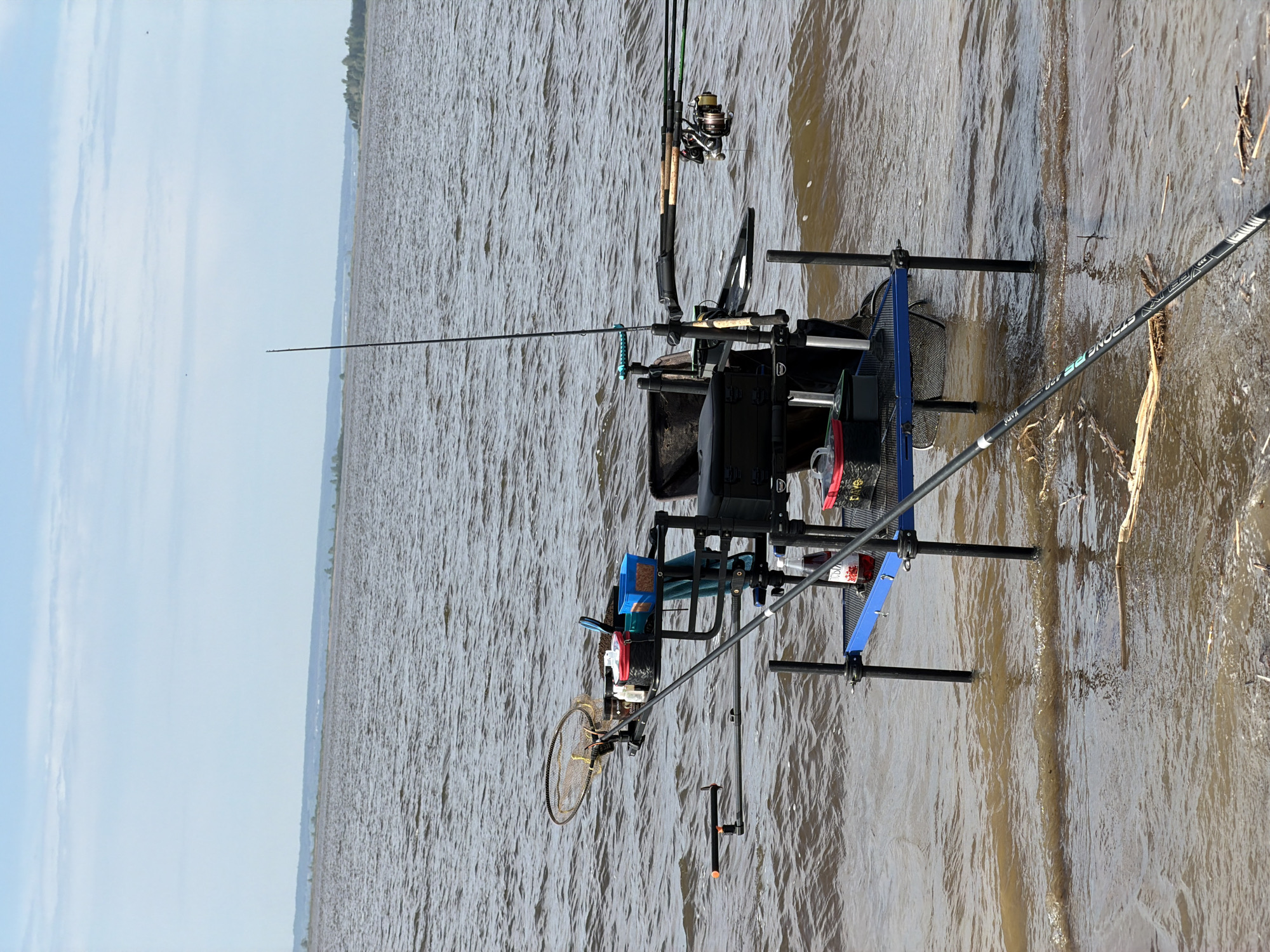 The image depicts fishing equipment set up near a body of water, with the shoreline visible in the foreground and a distant landmass across the water. The fishing setup includes multiple rods, reels, a tackle box, a net, and various accessories arranged on a blue-framed platform with adjustable legs submerged in the water. Sticks and debris are floating in the water near the shore. The sky is partly cloudy. The location is likely near Krasnyi Bor, Russia, based on the image's source.