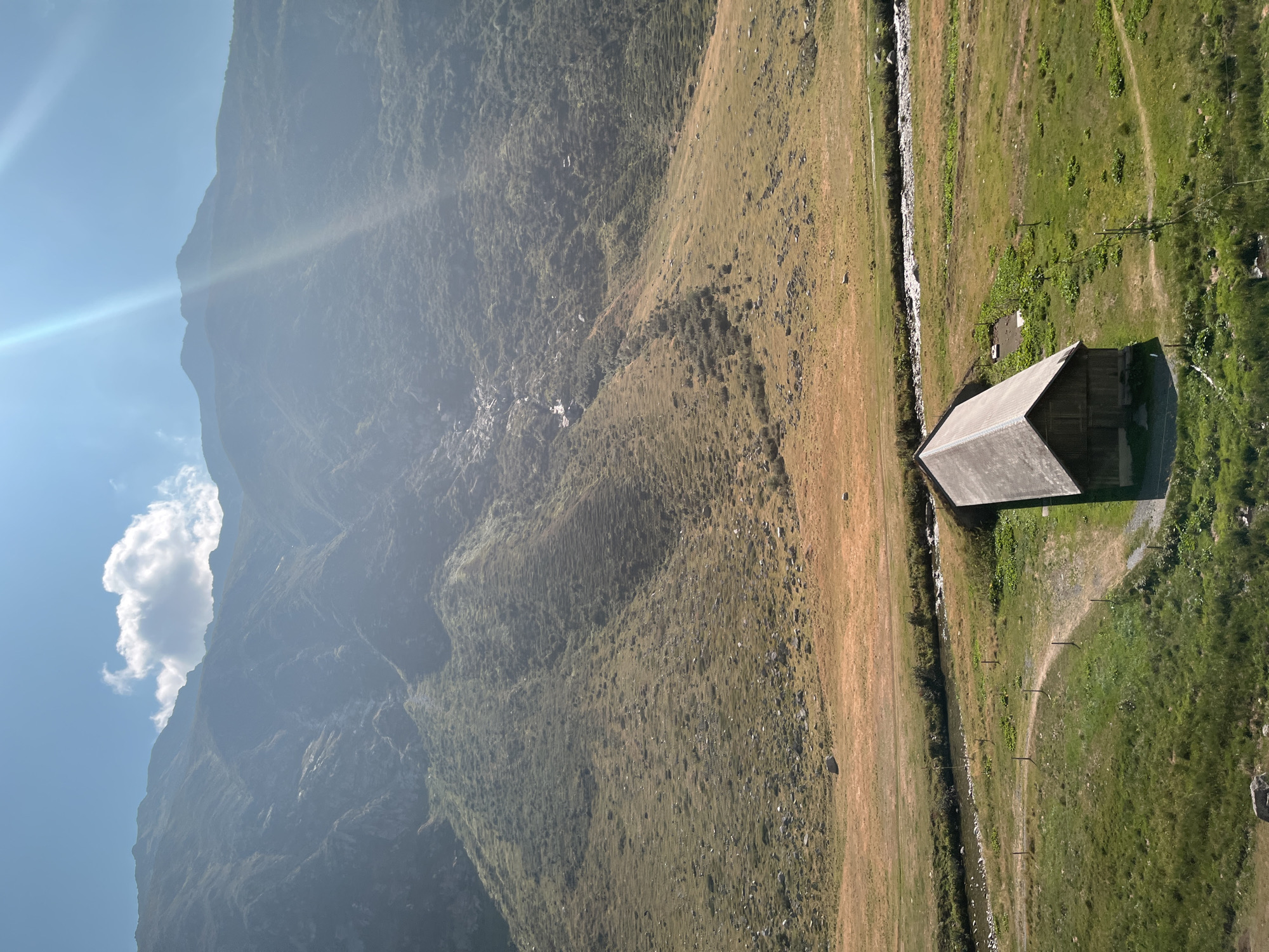 The image depicts a high-altitude rural landscape with a wooden barn nestled in a green meadow, situated in the vicinity of Hospental, Switzerland. The barn, with its steep, dark roof and wooden walls, is the primary man-made structure visible. A narrow, meandering dirt path leads to its entrance. A clear stream or river flows through the middle ground, separating the lower green fields from the higher, drier, golden-hued terrain. In the background, imposing mountains rise steeply, their slopes covered in sparse vegetation and rocky outcrops. The sky is a bright blue with a single, fluffy white cloud, and strong sunbeams create a lens flare effect in the upper right portion of the frame. The overall environment suggests a natural, remote mountain setting characteristic of the Swiss Alps.
