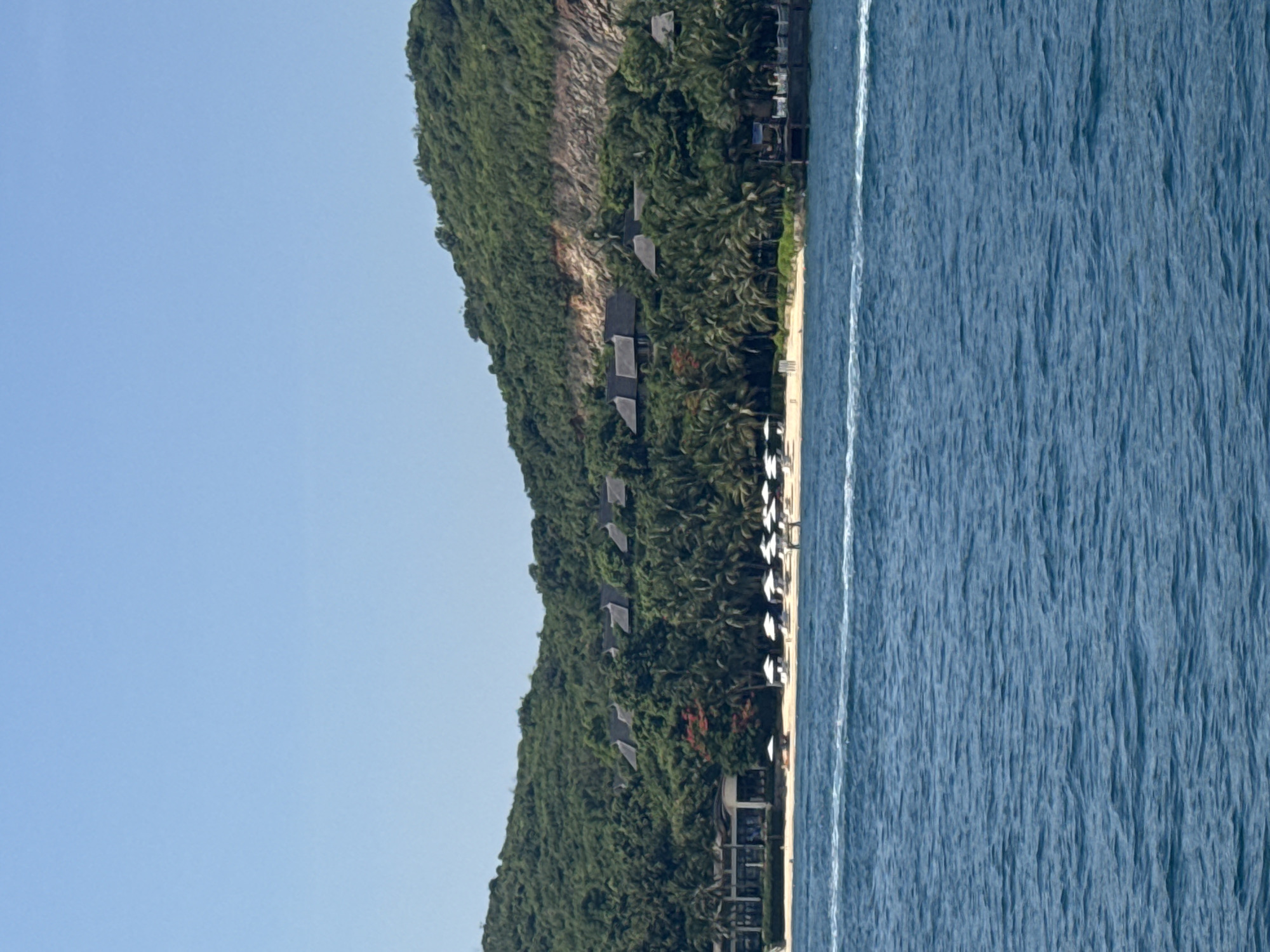This image shows a coastal resort located on a lush green hillside overlooking the ocean in Nha Trang Ward, Vietnam. The resort features buildings with dark, sloped roofs nestled among palm trees and dense vegetation. In the foreground, the blue ocean exhibits gentle waves, with a white wake suggesting a boat or jet ski has recently passed. Along the sandy beach, rows of white umbrellas and lounge chairs are visible, indicating a recreational area. A larger building complex with a modern architectural style is situated on the left side of the beach. The sky is clear and bright blue.