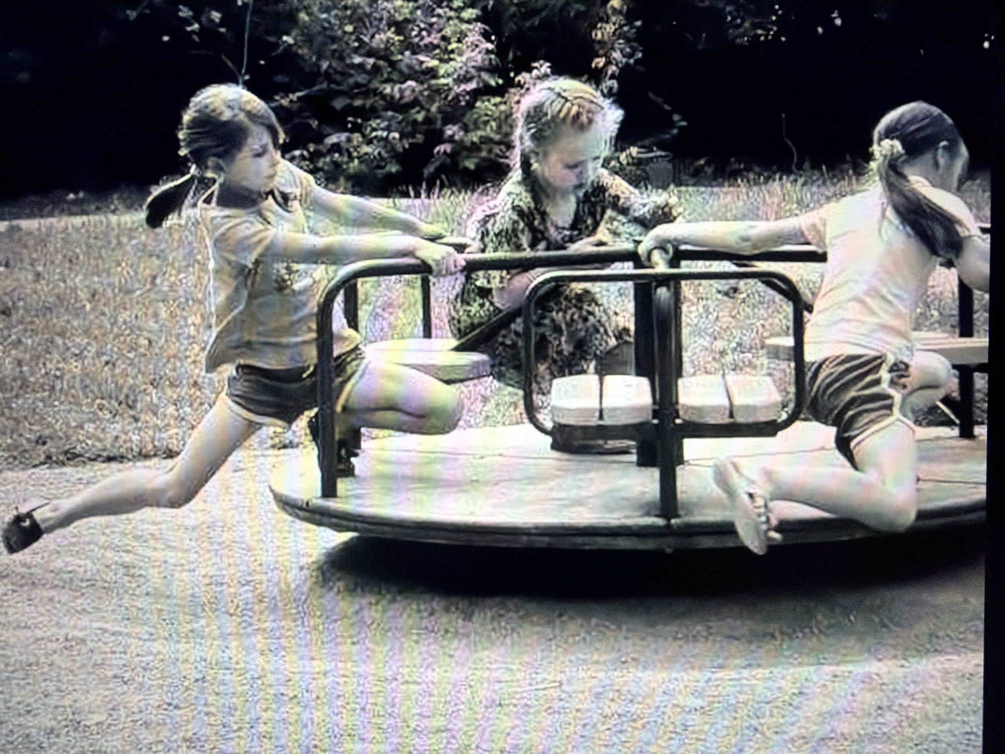 Three young children are on a merry-go-round in a grassy outdoor area with trees in the background. The child on the left, wearing a t-shirt and shorts, is leaning back with her legs extended to the side. The child in the middle, wearing a patterned dress, is seated and holding onto the merry-go-round's bars. The child on the right, wearing a t-shirt and shorts, is also holding onto the bars and is in a kneeling position. The merry-go-round has a wooden platform and black metal bars. The ground around the merry-go-round is covered in gravel and grass. The lighting suggests it is daytime.