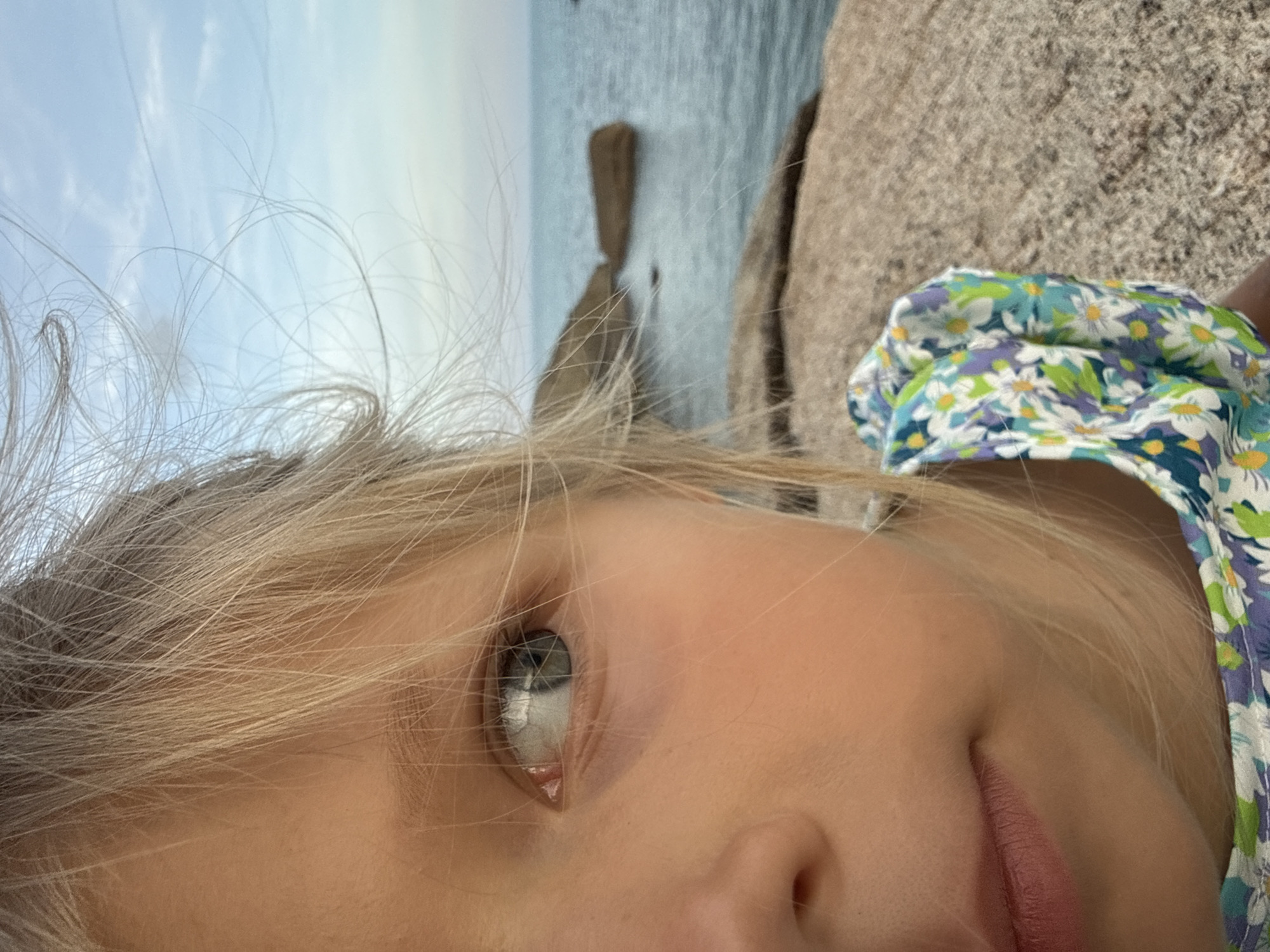 A close-up, half-face portrait of a young girl with blond hair is shown outdoors, with the sea and sky in the background. She is sitting on a large, rocky surface, possibly at a beach in Baan Hua Thanon, Thailand. Her hair is windswept, obscuring part of her face. The girl is wearing a floral-print dress with blue, green, and white patterns. Her left eye is clearly visible, and she is looking towards the left of the frame. The sea is calm and blue, with a few large rocks protruding from the water. The sky is a pale blue with light clouds. The overall impression is one of a natural, serene setting.