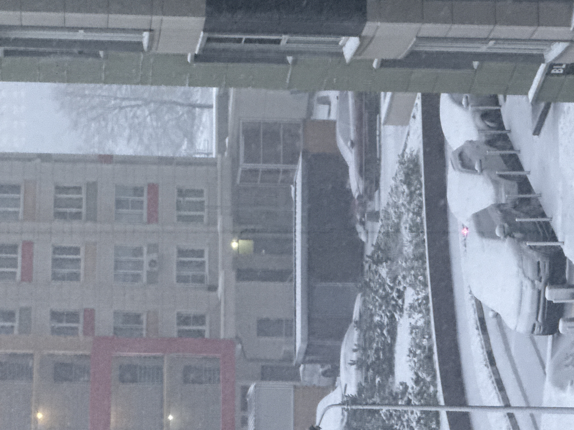 The image depicts a snowy scene in Khimki, Russia. A multi-story building with multiple windows and red accents on the facade is visible in the background. In the foreground, snow-covered vehicles line a street or parking area adjacent to a row of snow-laden bushes. Above this scene, part of a building overhang with visible architectural details. It appears to be snowing heavily, as evidenced by the blurred visibility and the thick snow accumulation.