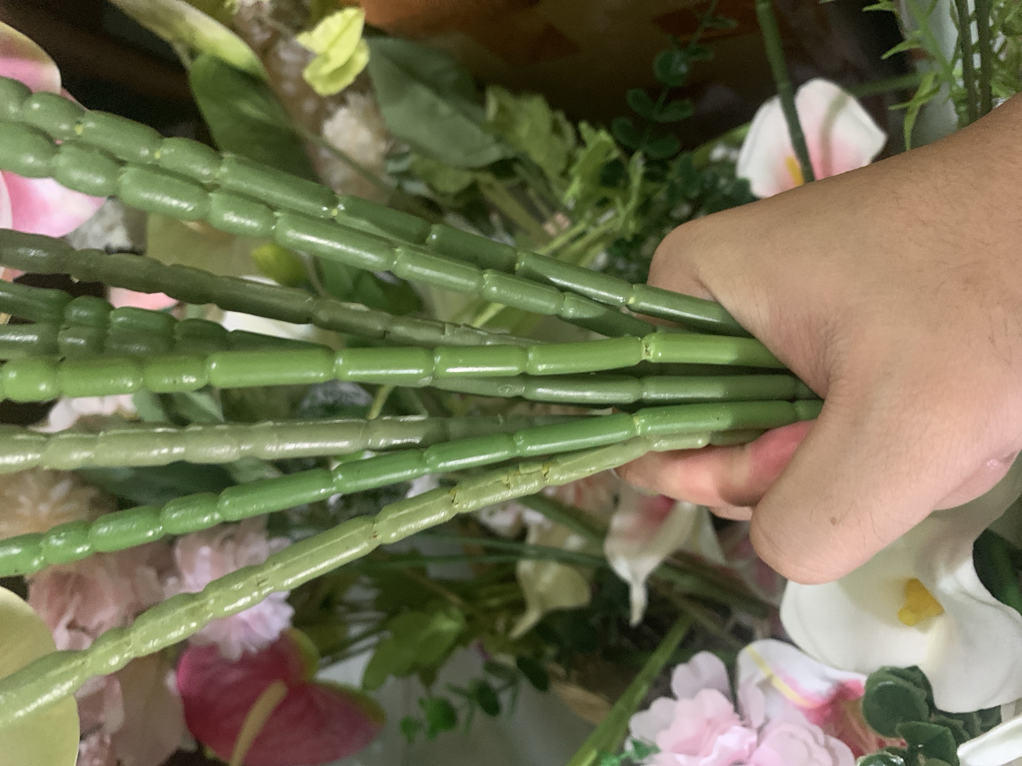 A person's hand is holding a bouquet of artificial flowers. The flowers are in various shades of pink and white, with green stems. The stems of some of the flowers are visible, and they are segmented and appear to be made of plastic. The background is blurred but appears to be an indoor setting with more flowers. The image was taken in Kuala Pembuang, Indonesia.