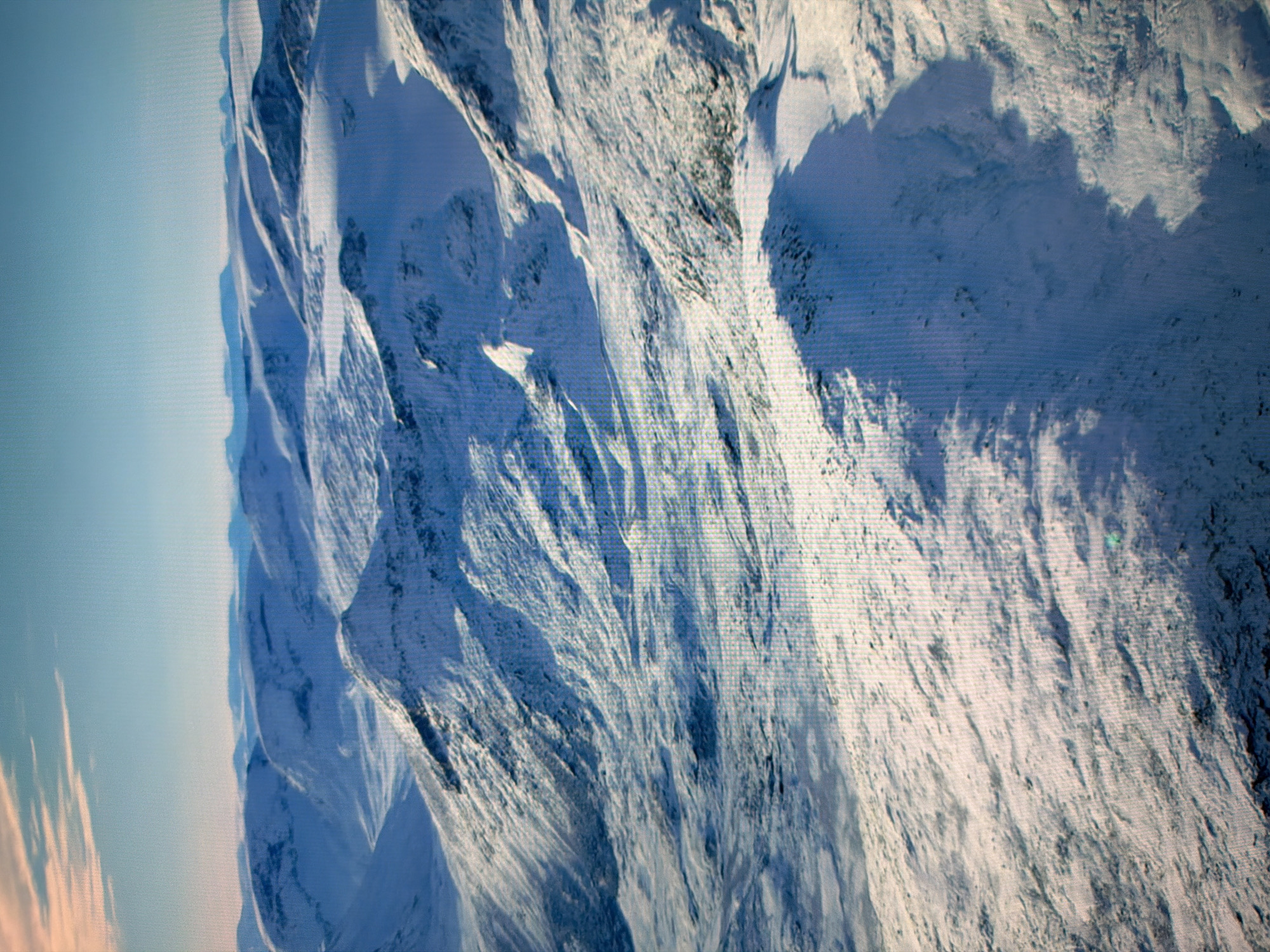 This image shows a panoramic view of a snow-covered mountain landscape under a clear blue sky with wispy clouds. The mountains are rolling and appear to be uninhabited. The terrain is characterized by gentle slopes and peaks, with shadows indicating a low sun angle, suggesting either early morning or late afternoon. The snow cover is extensive, with varying textures and depths creating patterns of light and shadow across the landscape. There is no indication of any urban environment, human presence, or activity in this image. Therefore, it does not depict any location in the city of Kyiv, Ukraine.