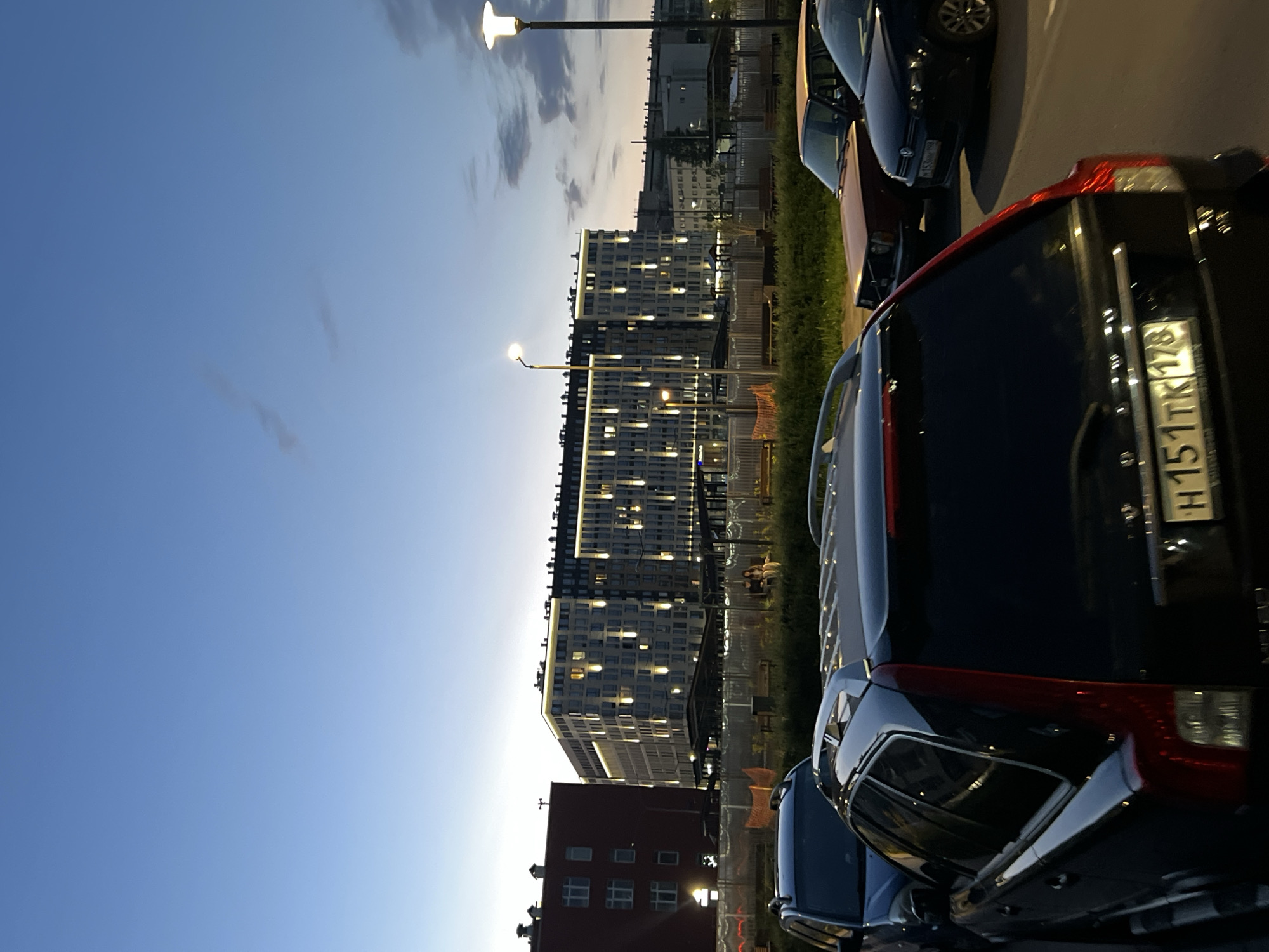 The image depicts an urban street scene at twilight, with the sky transitioning from light blue at the horizon to a deeper blue overhead, exhibiting scattered thin clouds. Artificial lights are active on buildings and streetlights.
In the foreground, a dark-colored Volvo V70 station wagon is parked, viewed from the rear, displaying Russian license plate "H151TK 178". To its immediate right, an older, reddish-brown sedan is visible. Further right, a dark Volkswagen Golf is parked, showing its front-left portion with a partially visible Russian license plate "B153OP 178". Part of another dark vehicle is visible to the left of the Volvo V70. The vehicles are on an asphalt surface.
Behind the parked cars, a fenced green space is visible, containing multiple wooden benches and some orange construction netting near the fence line. Two individuals are seated on a bench within this green area, positioned towards the center of the image. Two streetlights with white illumination are prominent; one on the left with a curved arm, and another taller, straight pole on the far right.
In the mid-ground, a multi-story, modern building with light-colored facade panels is illuminated with a patterned array of vertical and horizontal lights, alongside lights from individual windows. This structure appears to be approximately 10-12 stories tall. To the left of this modern building, a red-brick building of approximately 5-6 stories with visible windows is present. Further buildings are visible in the distant background, less distinctly.
The "178" regional code on the visible Russian license plates indicates the location is within the city of Saint Petersburg, Russia, or its immediate surrounding Leningrad Oblast.