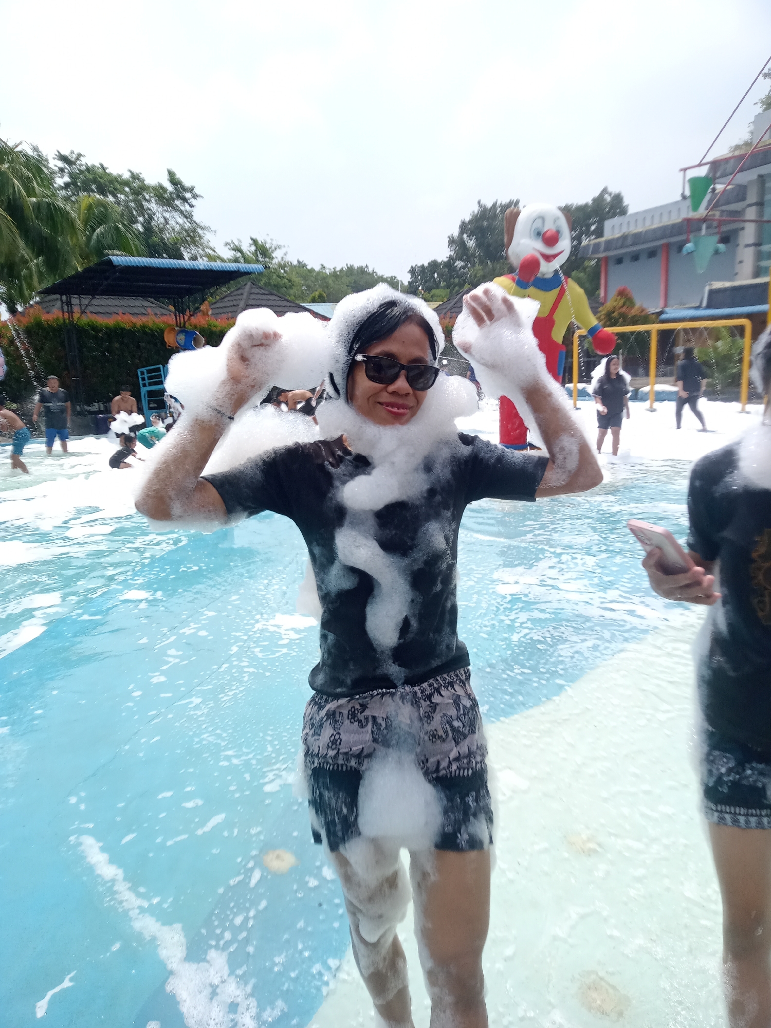 An outdoor water park, likely in the city of Baru, Indonesia, is depicted on a bright, overcast day. A woman, with black hair and wearing sunglasses and a dark t-shirt with a patterned skirt, is covered in foam, holding a handful of it up with her right hand. Her left hand is also covered in foam. In the background, a large clown inflatable stands prominently. Other people, including children and adults, are visible in the shallow pool, which is filled with foamy water. Trees and some park structures, including a shaded seating area and a yellow fence, are also present. The water appears blue and clear beneath the foam.