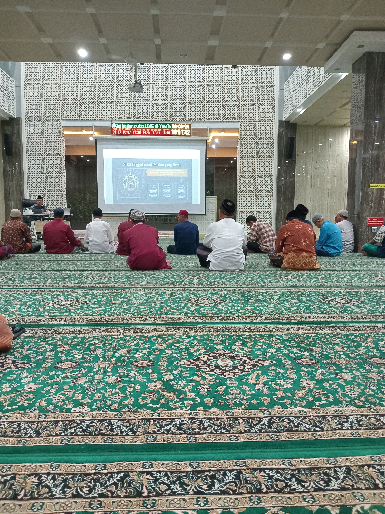 This is a medium shot, taken from a low angle, of a gathering of Muslim men sitting on a green carpet in what appears to be a mosque or a similar prayer hall. The men are facing away from the camera, towards a large projection screen displaying text in Indonesian, likely part of a presentation or lecture. The screen also shows a digital clock displaying prayer times. The architecture suggests a modern building with decorative latticework and marble columns. The men are dressed in traditional Islamic attire, with some wearing skullcaps. The overall atmosphere is one of communal gathering for religious or educational purposes. The specific location in Randuagung, Indonesia, is implied but not visually confirmed within the image itself.