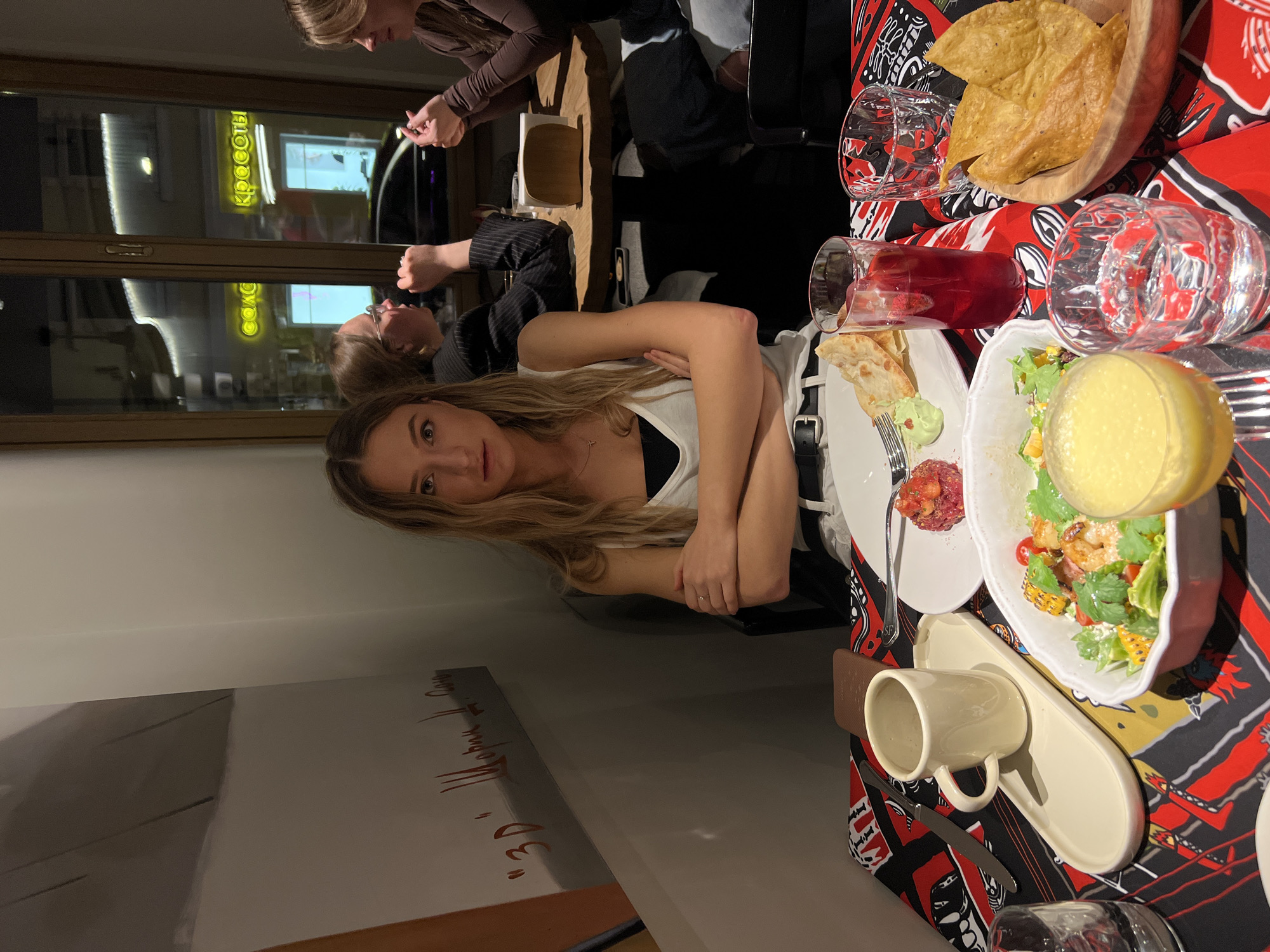 A young woman with long blond hair is sitting at a table in a restaurant. She has her arms crossed and is looking at the camera. On the table, there is a salad, a drink, and some tortillas. There are other people at the table, but they are out of focus. Through the window, the word "Красоты" can be seen, which translates to "Beauty" in Russian, which indicates that the location is in the city of Moscow, Russia.