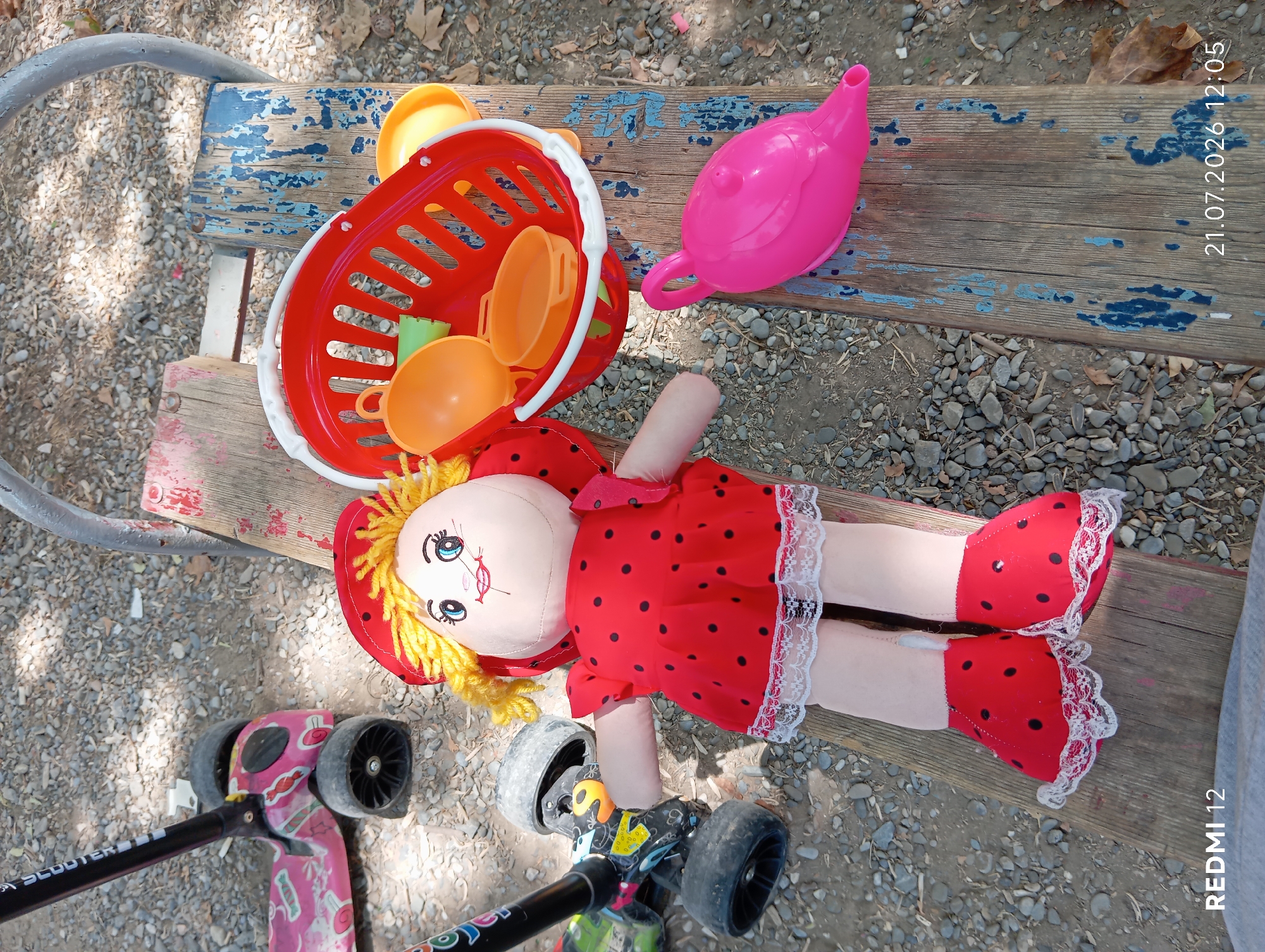 A doll dressed in a red and black polka dot outfit with lace trim lies on a wooden bench outdoors. Beside the doll, a red plastic basket contains toy kitchenware in orange and green. A pink plastic watering can is also on the bench. In the background, two scooters are partially visible, one pink and one black and green. The scene is set on a gravelly surface, possibly in a park or playground in Bekabad, Uzbekistan. The lighting suggests it is daytime.