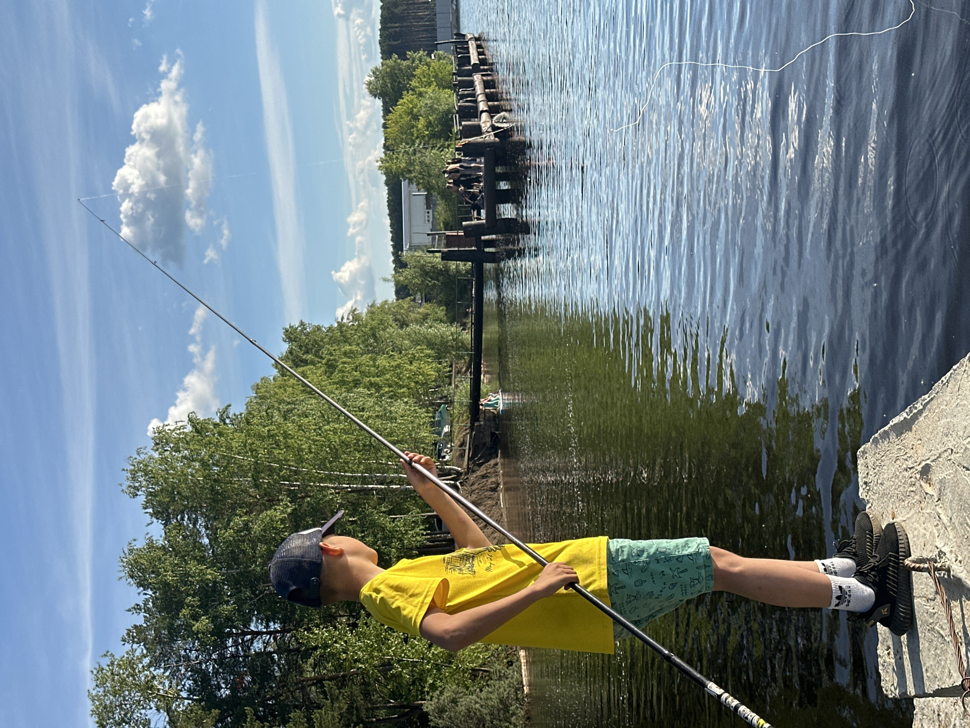 In the city of Chaikovsky, Russia, on a sunny day with scattered clouds, a young boy, facing away from the camera, stands on a concrete pier and fishes in a body of water. He wears a yellow t-shirt, light green shorts with a pattern of palm trees and waves, white socks with black stripes, and black athletic shoes. He holds a fishing rod, casting a line into the water. In the background, across the water, is a wooden pier with several people standing and sitting on it. Beyond the pier, trees line the shore, and in the distance, a white building is visible. The water is calm with ripples.