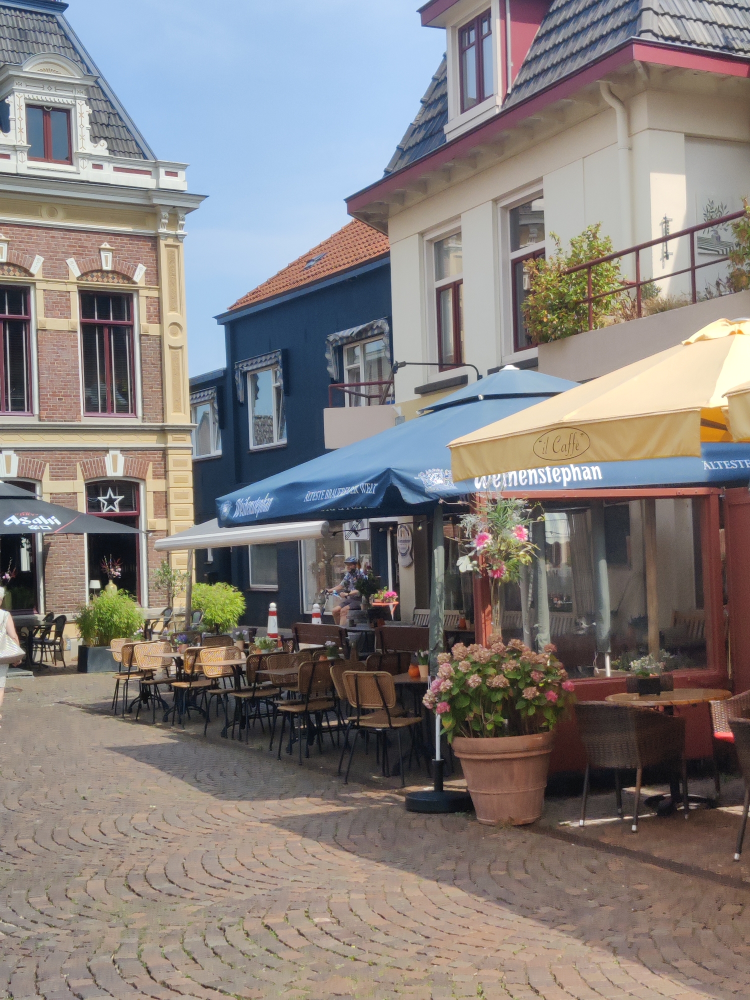 The image depicts a street scene in Winterswijk, Netherlands, featuring the exterior of a cafe with outdoor seating. The cafe has a light-colored facade and a large awning displaying the text "weissenstephan" and "il Caffè." Several tables and chairs are arranged on a cobblestone patio, some covered by umbrellas. A large potted plant with pink flowers is prominently displayed near the entrance. In the background, other buildings with distinctive Dutch architecture are visible, including a blue building and a building with red brick and decorative trim. The sky is clear and blue. There is one person partially visible on the left side of the image, walking away from the camera. Another person is visible through the cafe window, seated at a table.