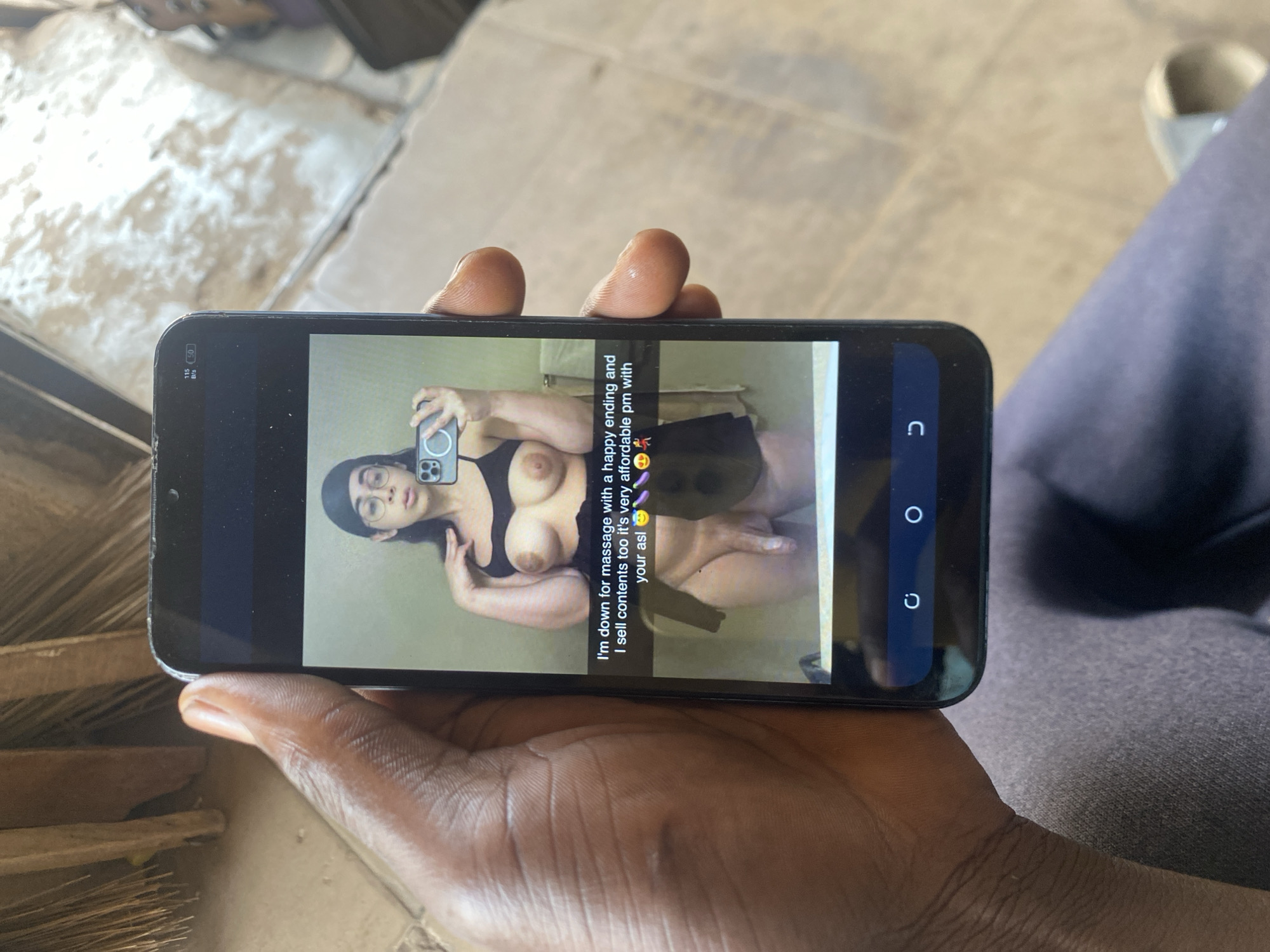 A dark-skinned individual is holding a smartphone in their hand, displaying a photograph of a woman in lingerie with a sexually suggestive caption. The phone's screen reveals a selfie-style image of the woman wearing glasses, a beanie, and lingerie, with text overlayed that reads, "I'm down for massage with a happy ending and I sell contents too it's very affordable pm with your asl." The background around the person holding the phone appears to be outdoors, possibly a residential area, with a combination of wood or straw materials and what seems like a concrete or tiled surface. The location of Bukuru, Nigeria is inferred from the context, but not directly visible in the image.